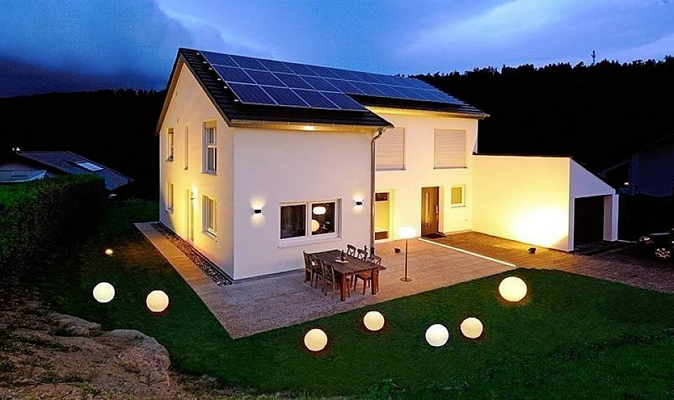 Zweistöckiges, modernes Haus bei Dämmerung, ausgestattet mit Solarzellen auf dem Dach. Beleuchteter Garten mit runden Lampen.