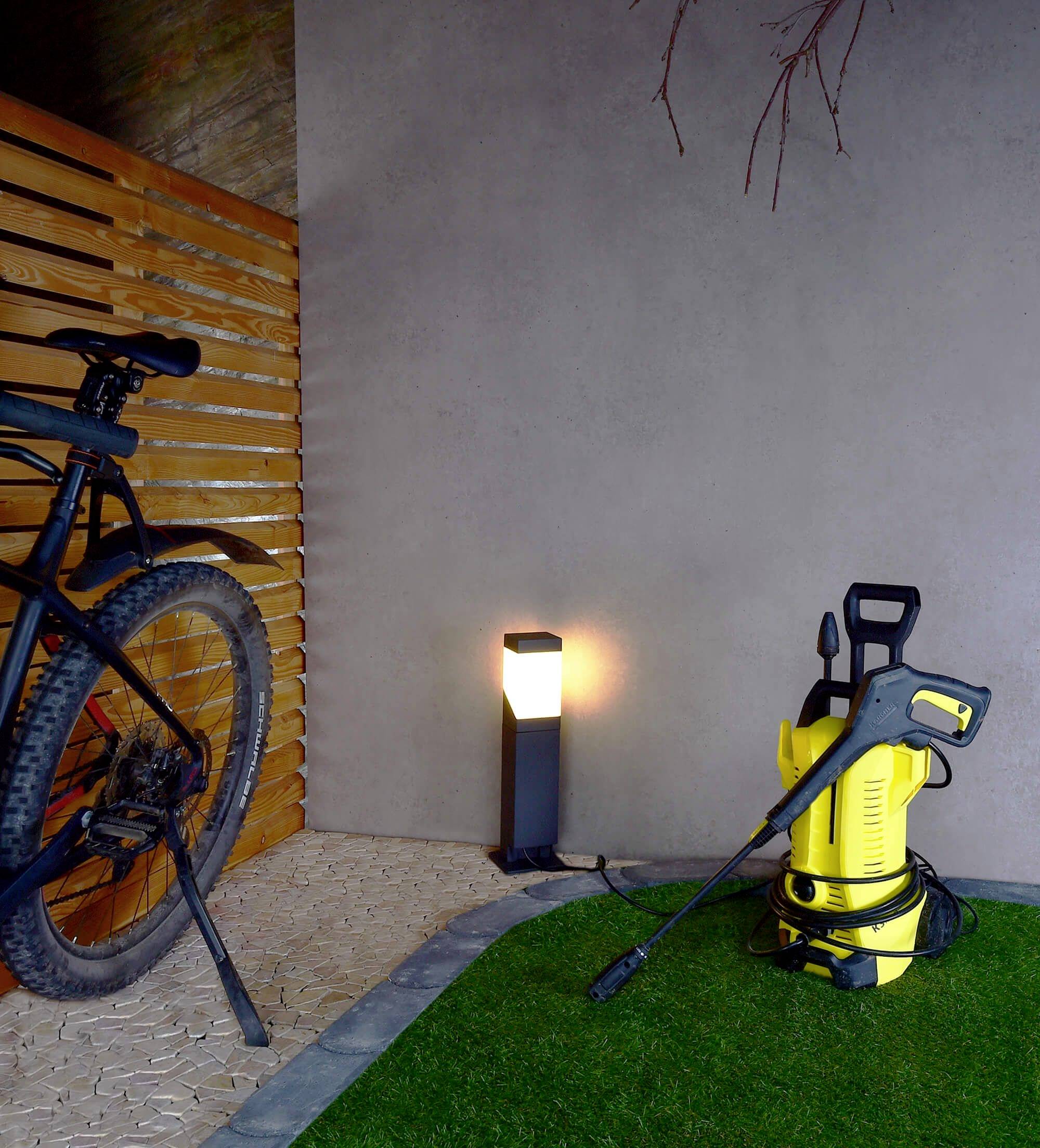 Ein gelbes Hochdruckreinigungsgerät steht neben einem Fahrrad auf künstlichem Rasen, daneben eine beleuchtete Lampe an Holzwand.