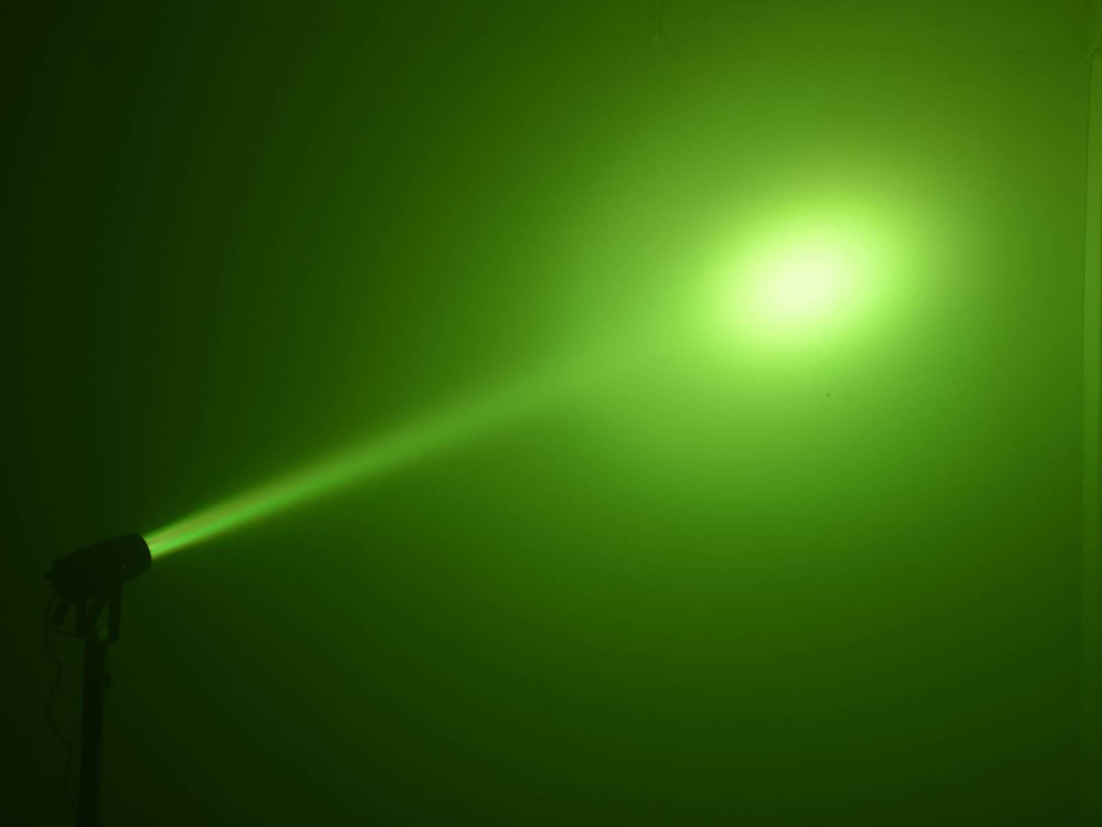 Grüner Laserstrahl leuchtet von links nach rechts durch einen dunklen Raum, bildet einen leuchtenden Punkt an der Wand.
