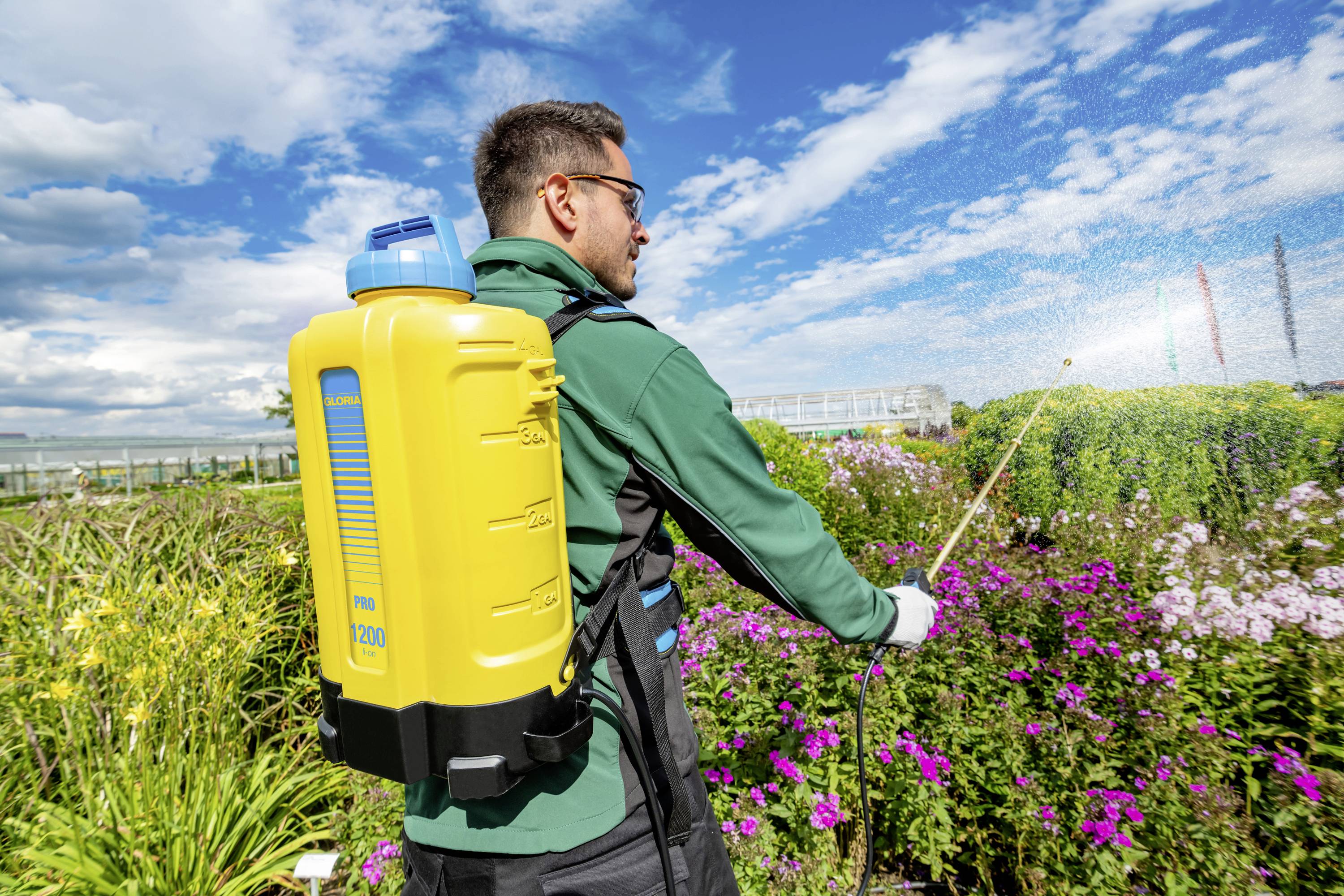 Ein Mann mit einer gelben Sprühvorrichtung auf dem Rücken bewässert Pflanzen in einem Garten unter blauem Himmel.