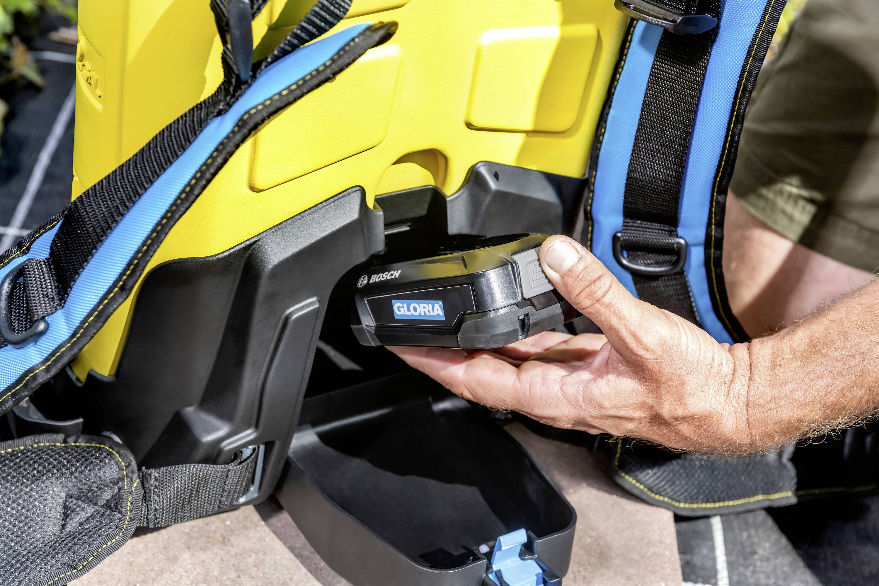 Eine Person legt einen Akku in ein gelbes elektrisches Gartensprühgerät mit blauen Riemen ein. Gerät ist von Marke 'GLORIA'.