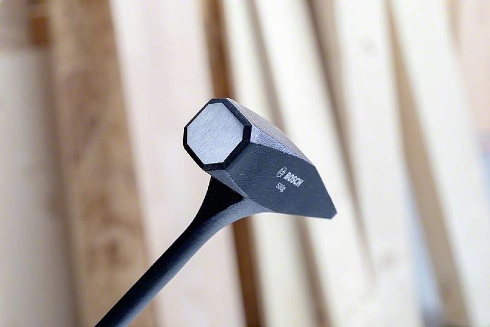 Ein Zimmermannshammer mit quadratischem Kopf und spitzem Ende vor unscharfem Holz im Hintergrund. Markenname 'BOSCH' sichtbar.