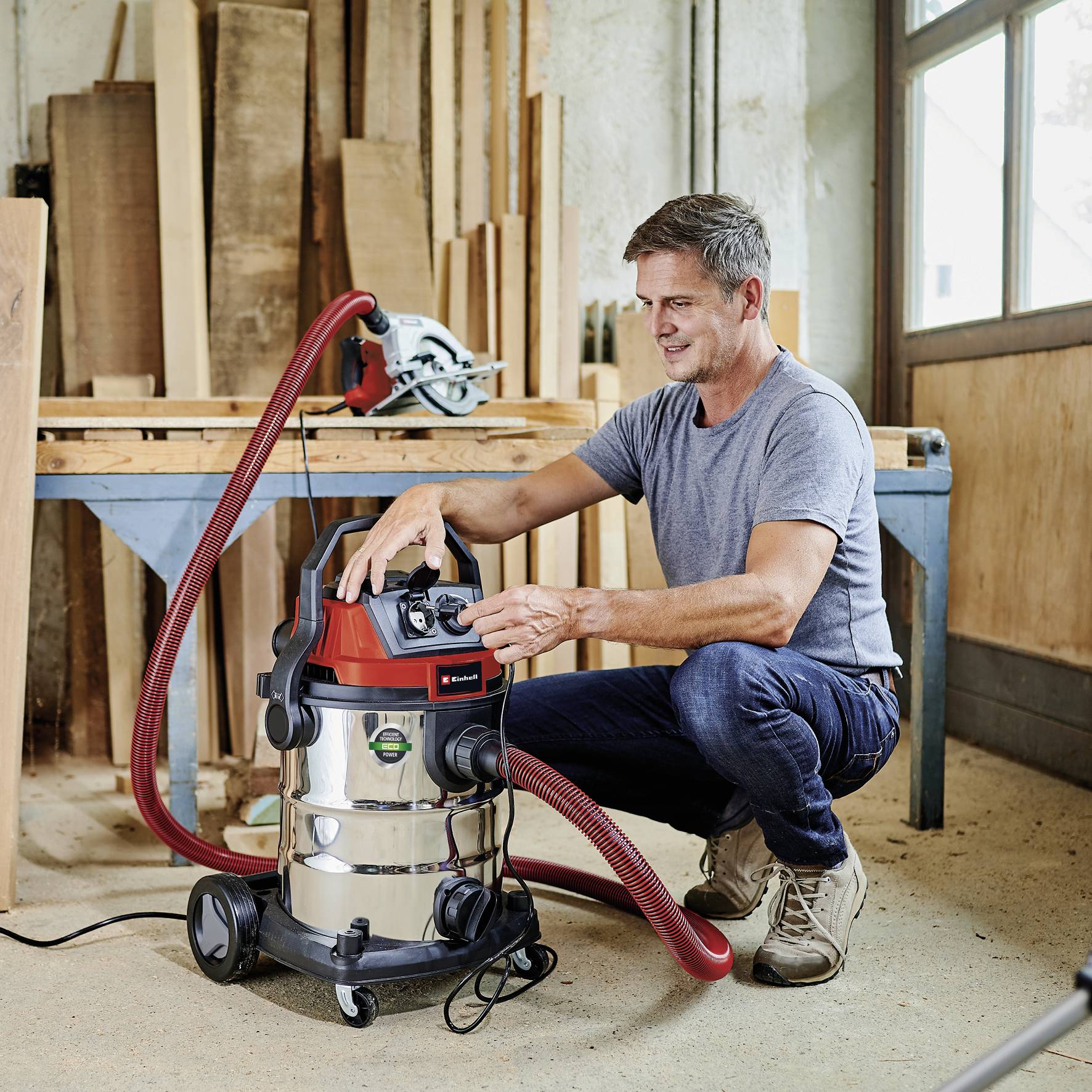 'Mann in Werkstatt benutzt Nass-Trockensauger neben Werkbank; im Hintergrund liegt eine Säge.'
