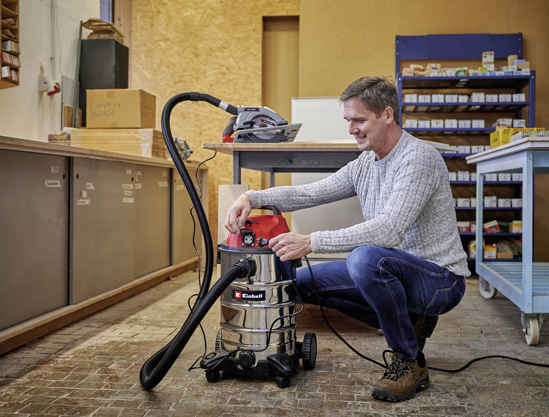 Ein Mann im Pullover überprüft einen Industriestaubsauger in einer Werkstattumgebung. Im Hintergrund sind Regale mit Werkzeugen zu sehen.