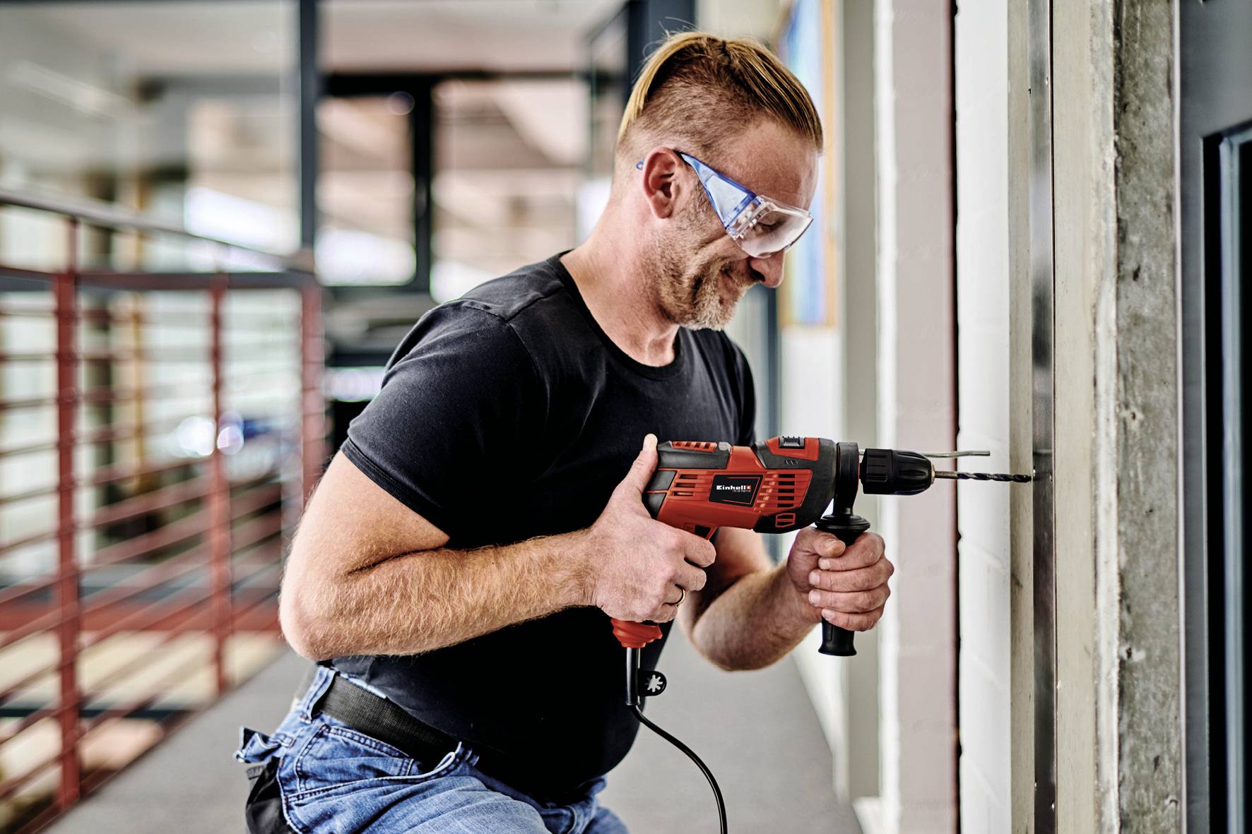 Ein Mann, der Schutzbrille trägt, bohrt mit einer roten Bohrmaschine in eine Wand. Er trägt ein schwarzes T-Shirt und Jeans.