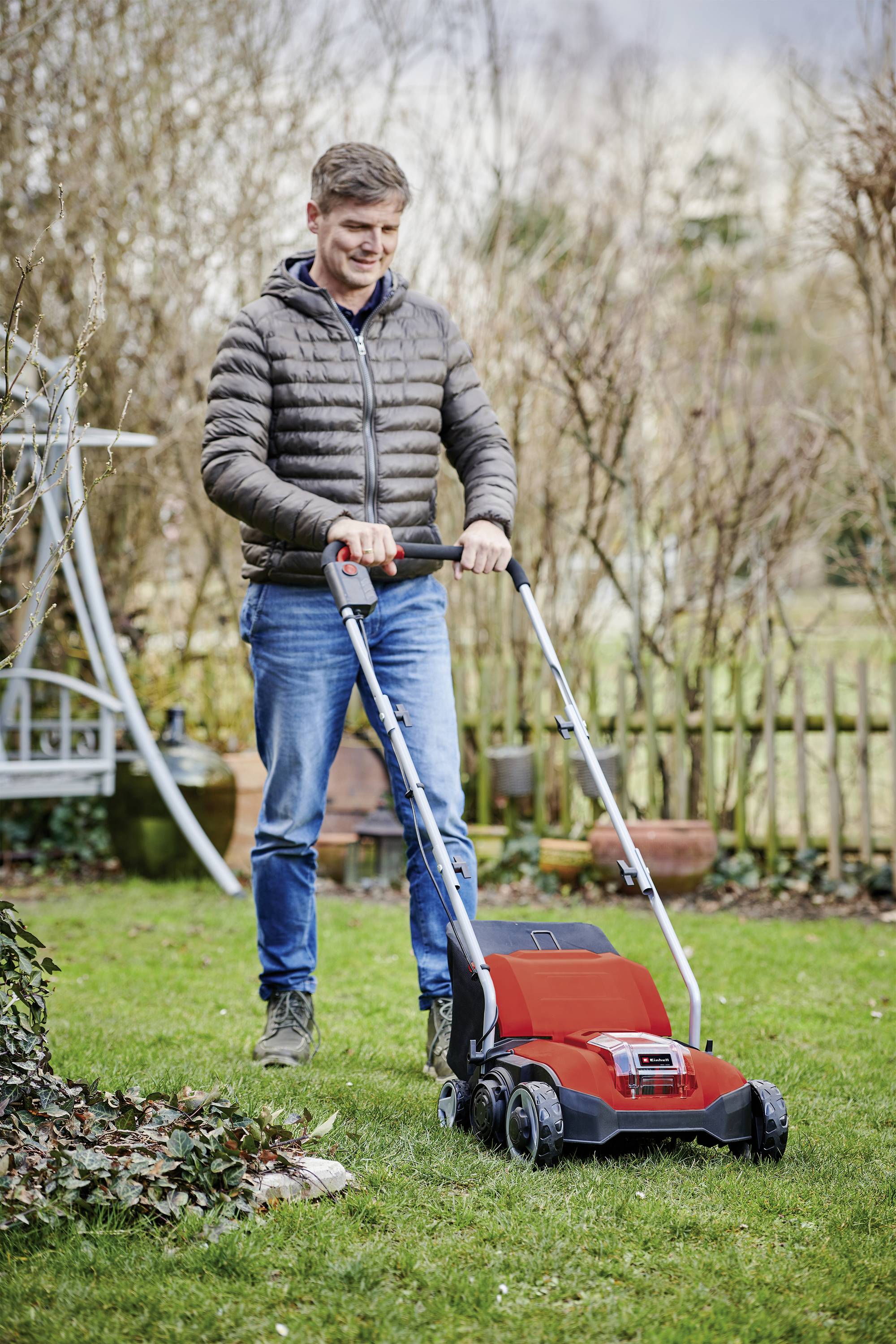Ein Mann in Freizeitkleidung schiebt einen roten Rasenmäher über einen grünen Rasen in einem Garten mit Holzzaun und kahlen Sträuchern.