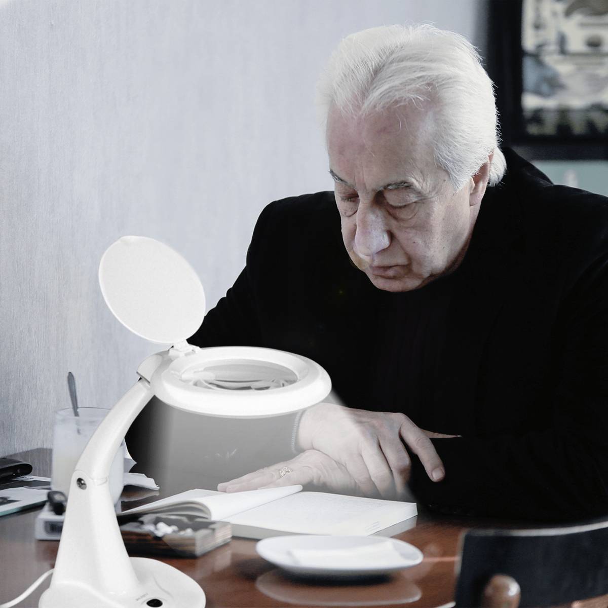 Älterer Mann mit weißem Haar liest an einem Tisch unter einer Schreibtischlampe, neben ihm liegt ein Notizbuch.