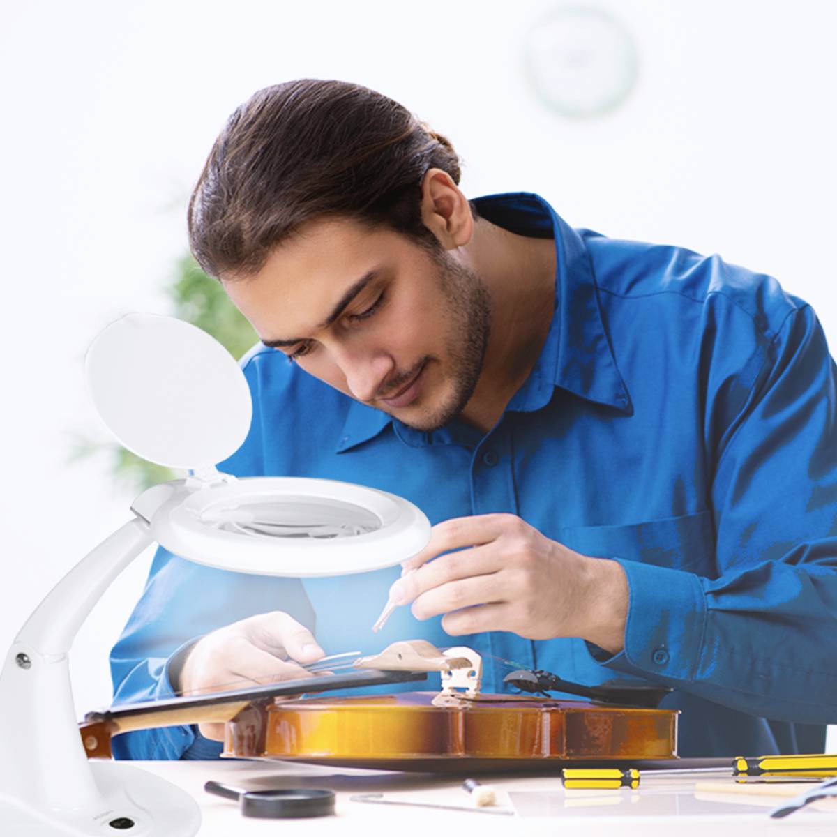 Ein Mann repariert eine Geige unter einer Vergrößerungslampe, konzentriert auf die Arbeit mit Werkzeugen auf dem Tisch.