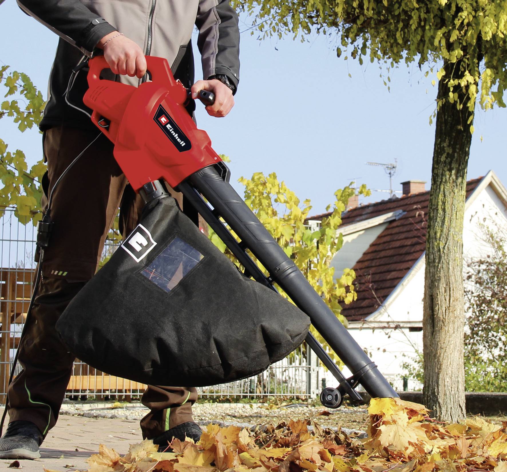 Eine Person verwendet einen roten Laubsauger auf einem Gehweg. Der Laubsauger saugt Blätter in einen schwarzen Beutel auf. Im Hintergrund sind Bäume und ein Haus erkennbar.