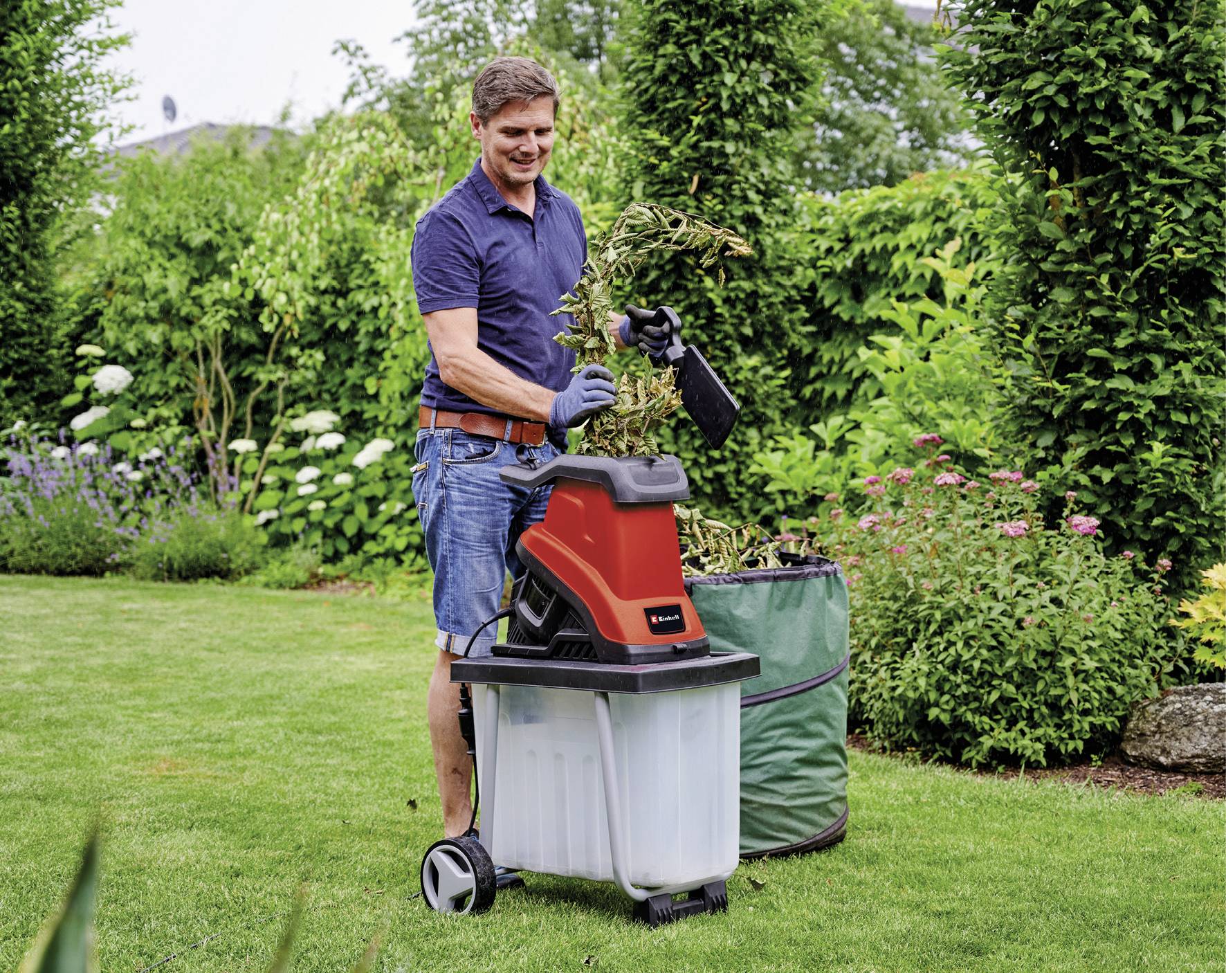 Ein Mann zerkleinert Pflanzenreste im Garten mit einem roten Gartenhäcksler. Im Hintergrund sind gepflegte Pflanzen und ein großer Laubsack zu sehen.