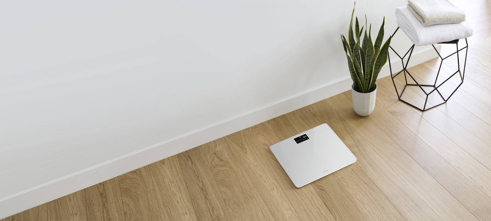 Waage auf Holzboden neben grünem Pflanzentopf und gefalteten Handtüchern, minimalistisches Badezimmerdekor.