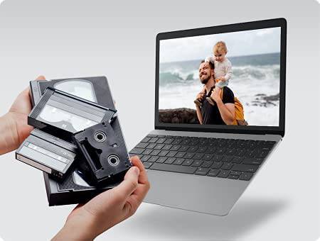Zu sehen sind zwei Hände, die alte Videokassetten halten, und ein Laptop, auf dem ein Foto eines Mannes mit Kind am Meer angezeigt wird.