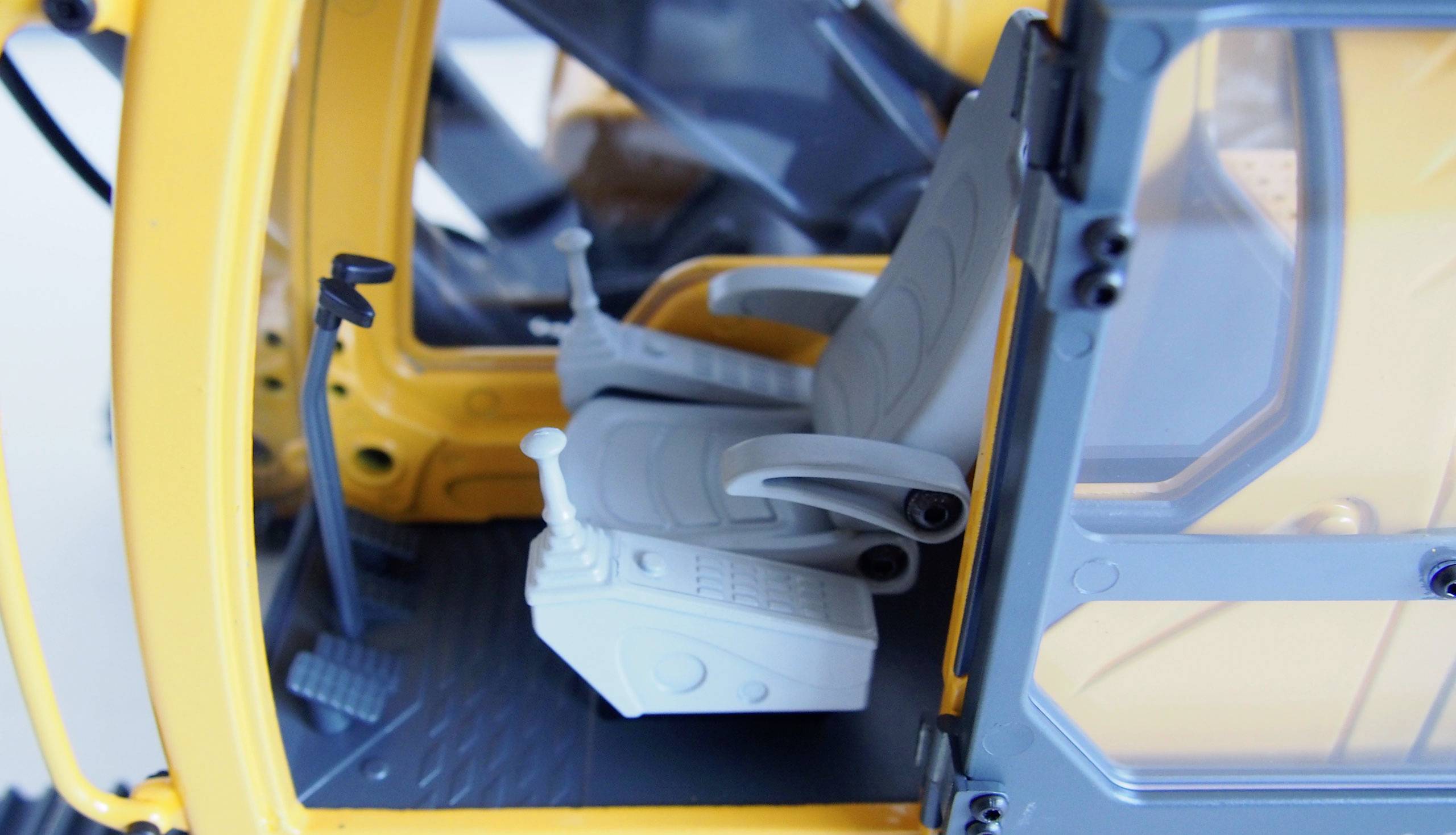 Innenraum einer gelben Baustellenmaschine mit zwei grauen Fahrersitzen und Bedienelementen. Sicht auf das Cockpit und die Steuerhebel.