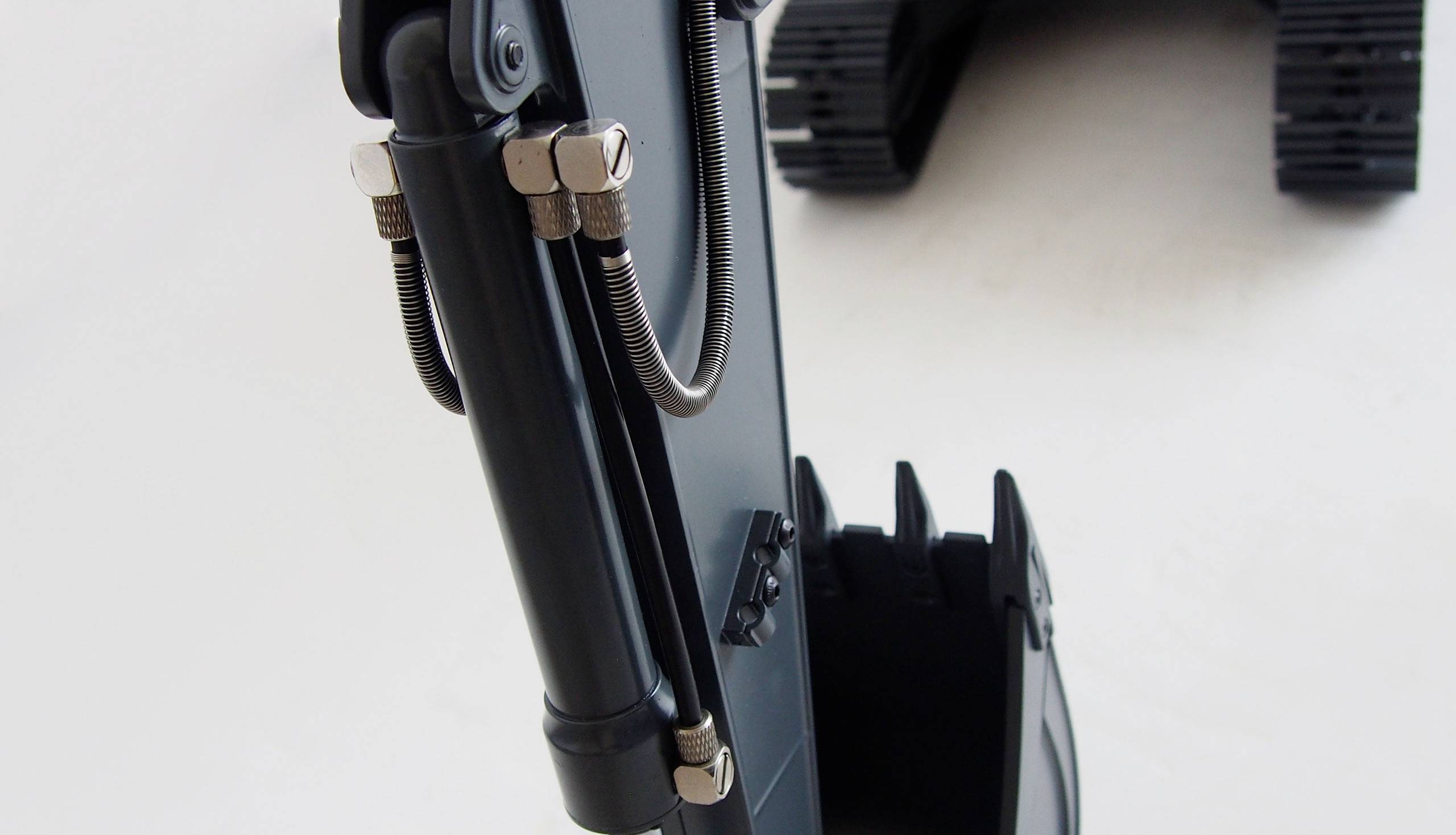 Nahaufnahme eines schwarzen Baggerarms mit Schaufel und Hydraulikschläuchen; Teile von Raupen sind sichtbar im Hintergrund.