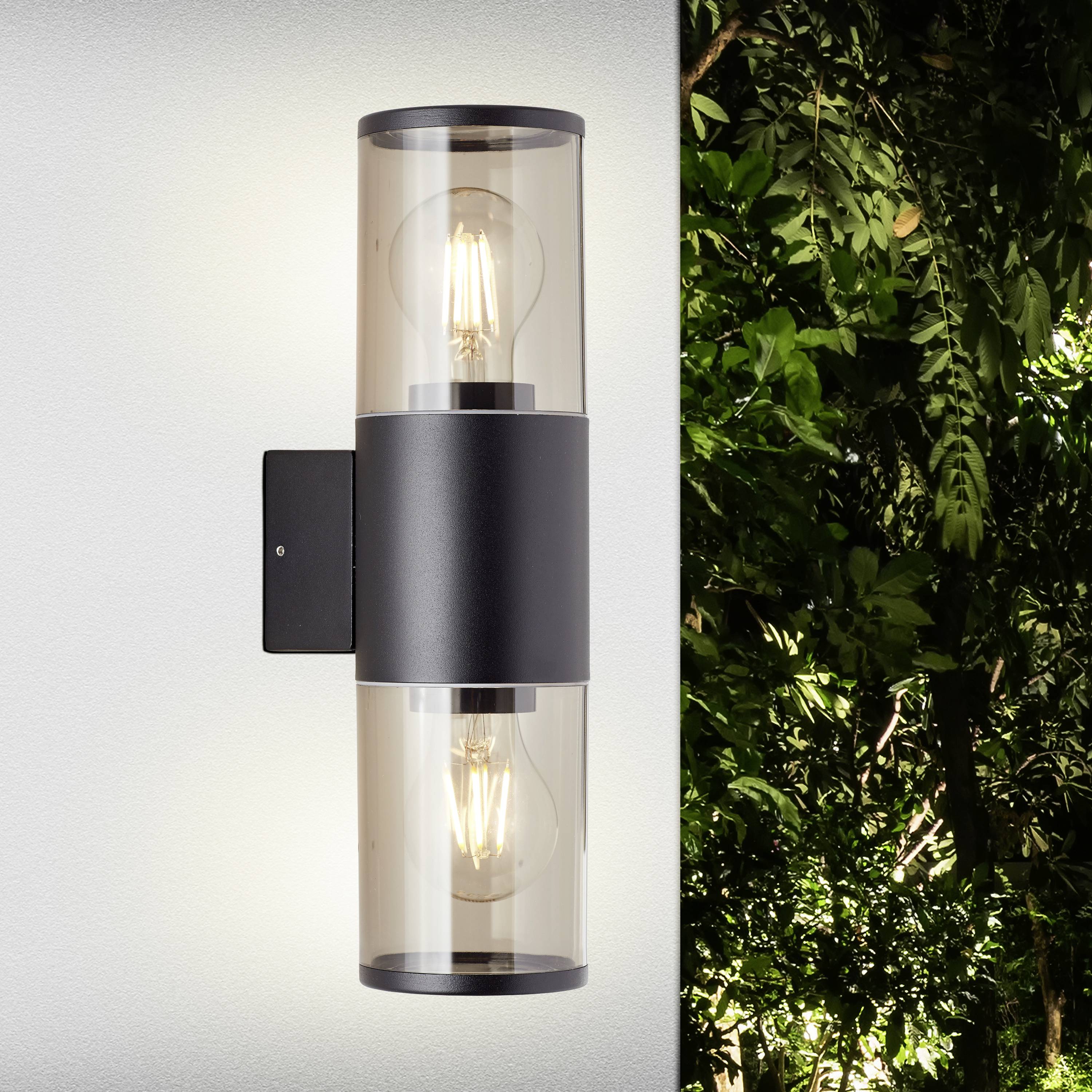 Moderne Außenwandlampe mit zwei Glühbirnen, montiert an einer weißen Wand. Rechts daneben grünes Laub im Hintergrund.