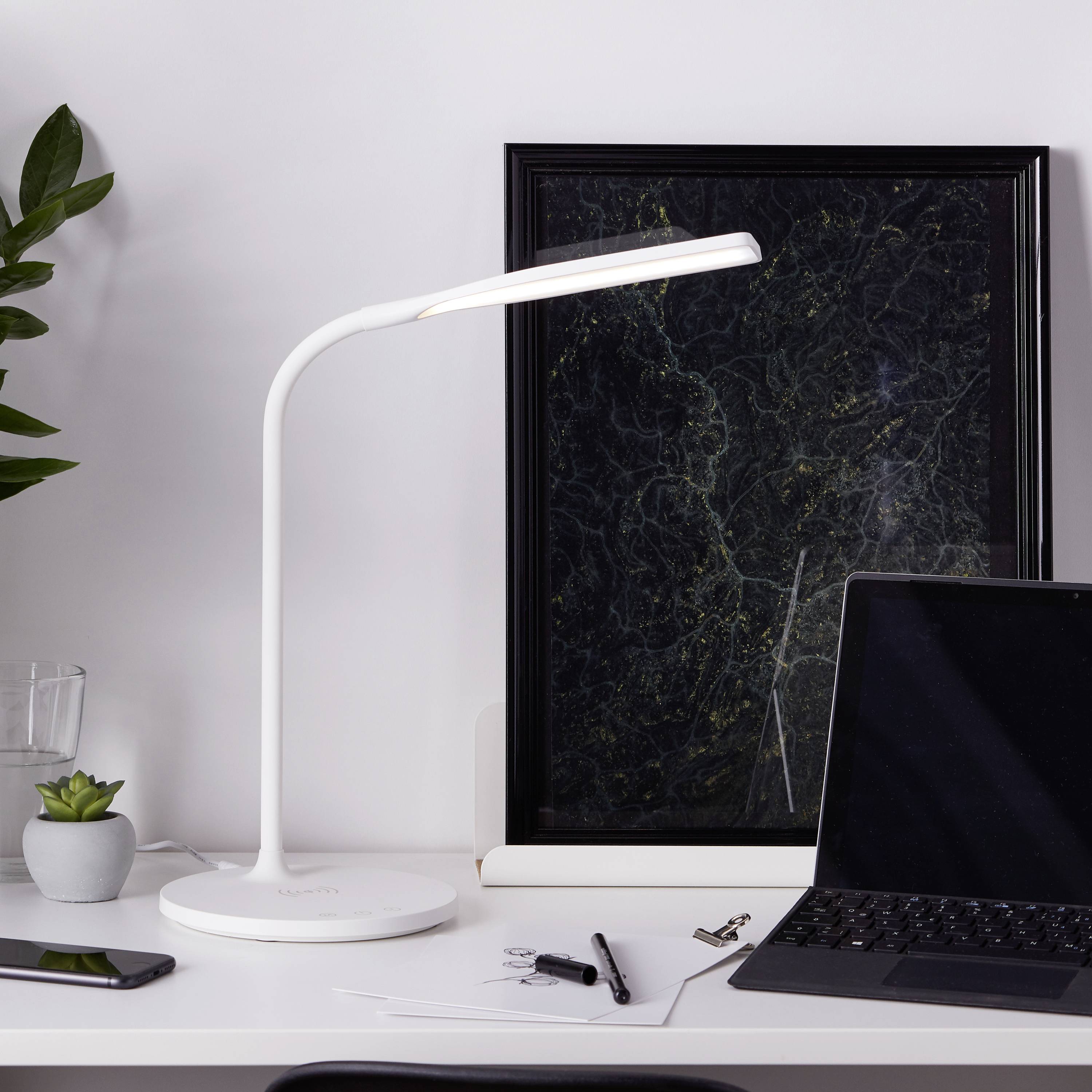 Schreibtisch mit moderner weißer Lampe, Laptop, Notizbuch, Stift und kleinen Pflanzen. Entspannende, minimalistische Arbeitsumgebung.