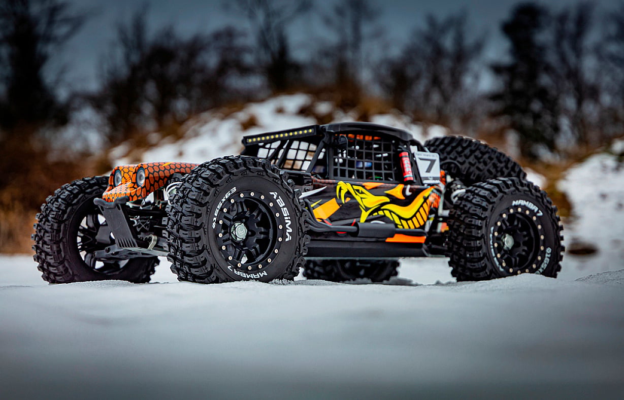 Ferngesteuertes Auto im Schnee, schwarz-orange mit auffälligem Grafikdesign an den Seiten. Grobe Reifen und schneebedeckter Hintergrund.