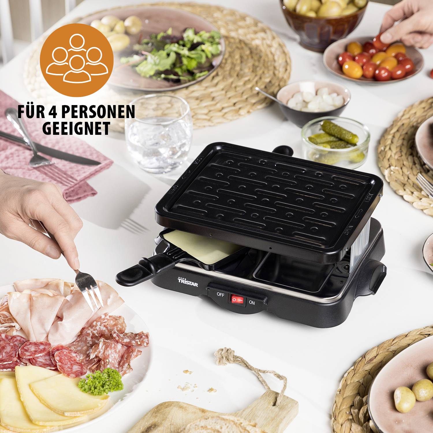 Ein Raclette-Grill auf einem Tisch, bereit für vier Personen. Es gibt Salat, eingelegte Gurken, eine Auswahl an Fleisch und Käse.