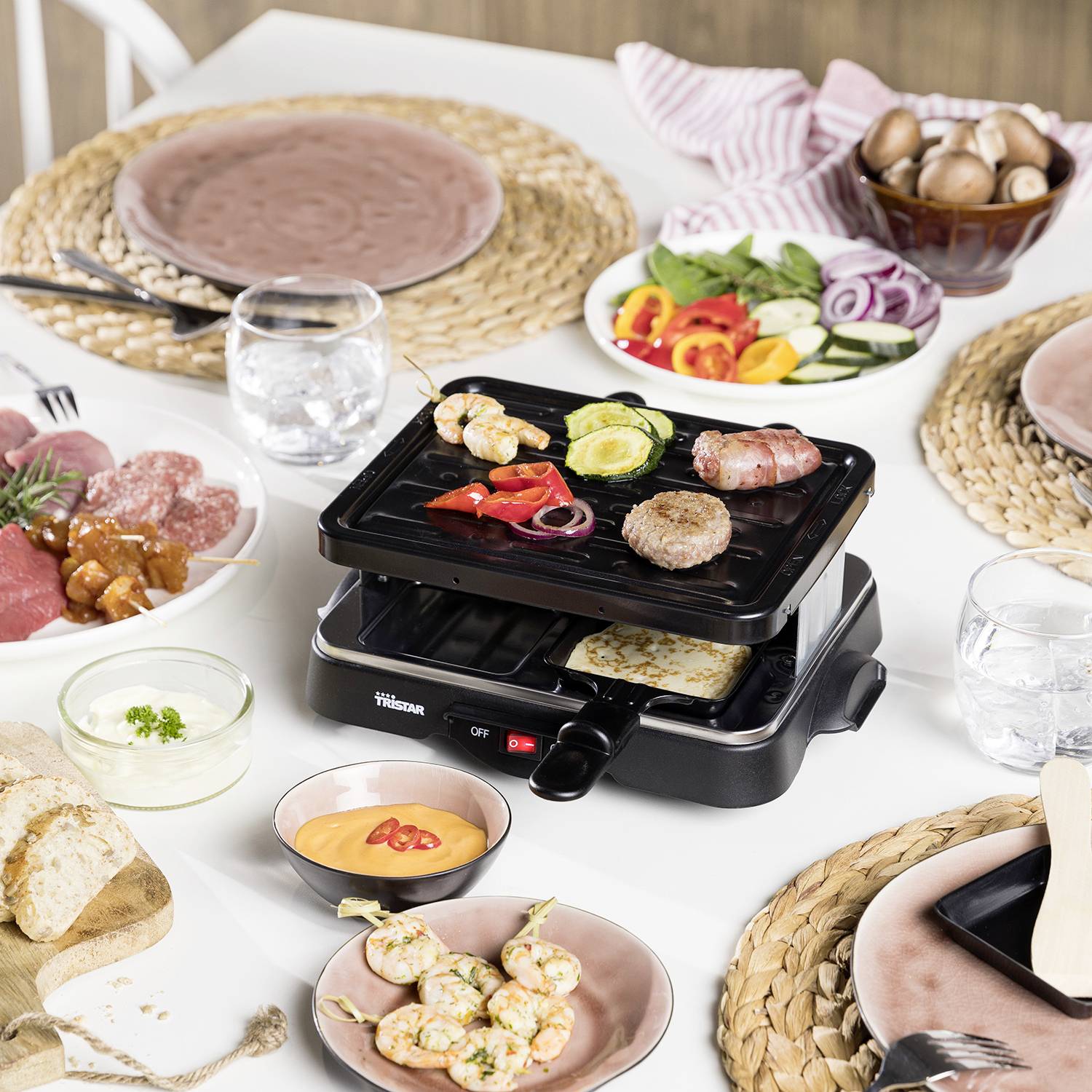 Ein Tisch ist gedeckt mit einem Raclette-Grill in der Mitte, umgeben von Tellern, Fleisch, Gemüse, Brot, Dips und Eiern.