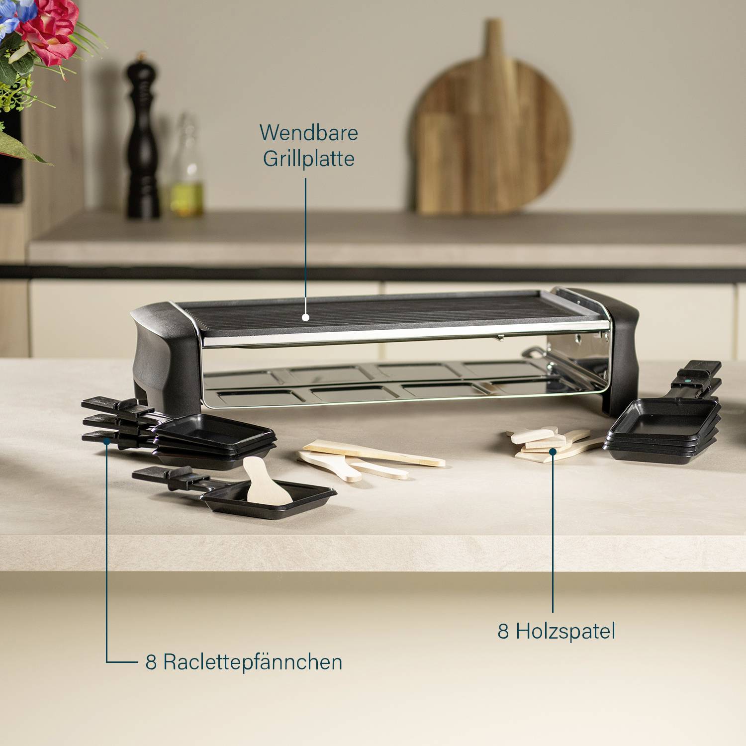 'Raclette-Grill mit Wendeplatte und 8 Pfännchen auf einer Küchenarbeitsplatte. Daneben 8 Holzspatel. Im Hintergrund Dekoration.'