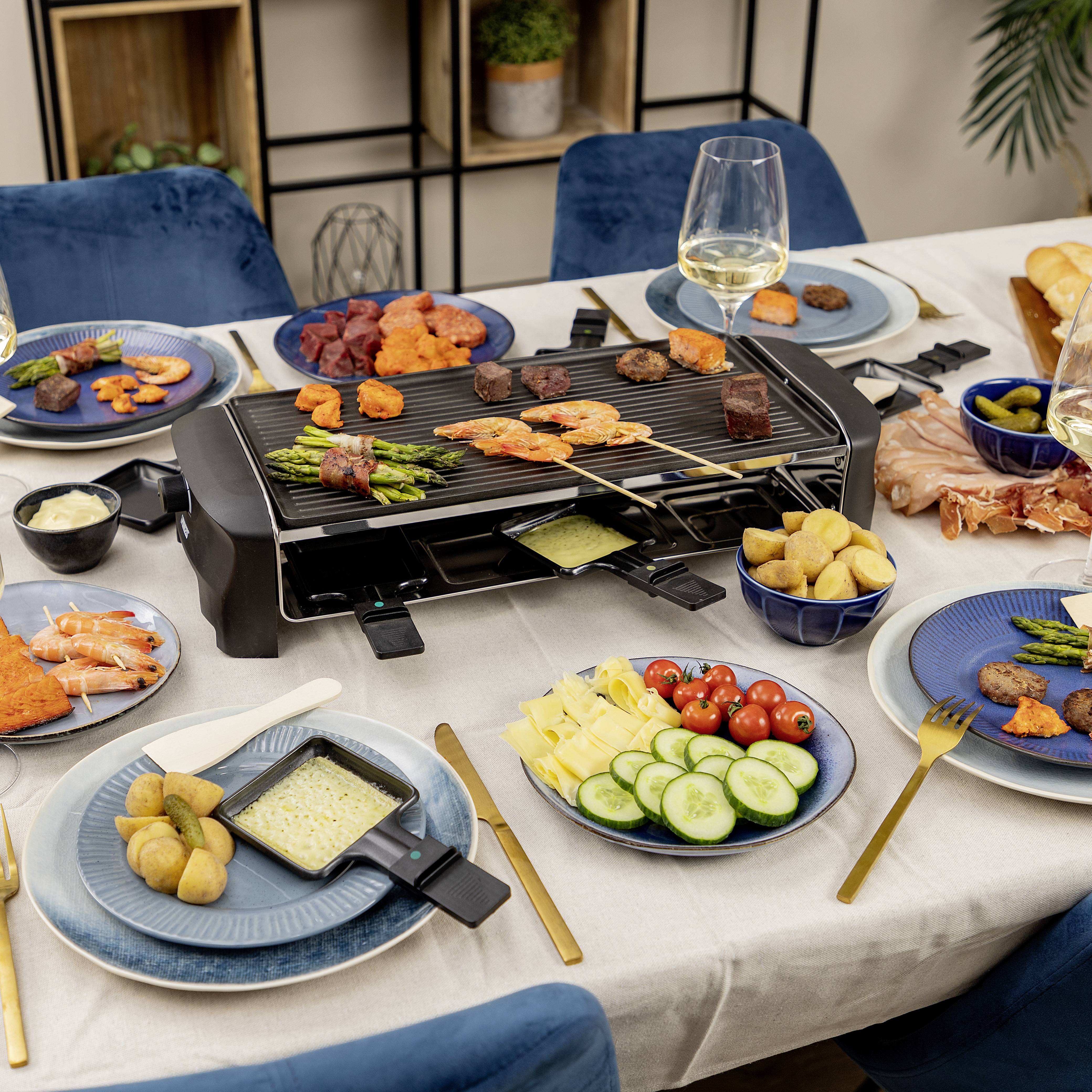 Ein Tisch mit Raclette-Grill, darauf Fleisch und Gemüse. Auf Tellern liegen Kartoffeln, Salat und Saucen. Gläser mit Weißwein stehen daneben.