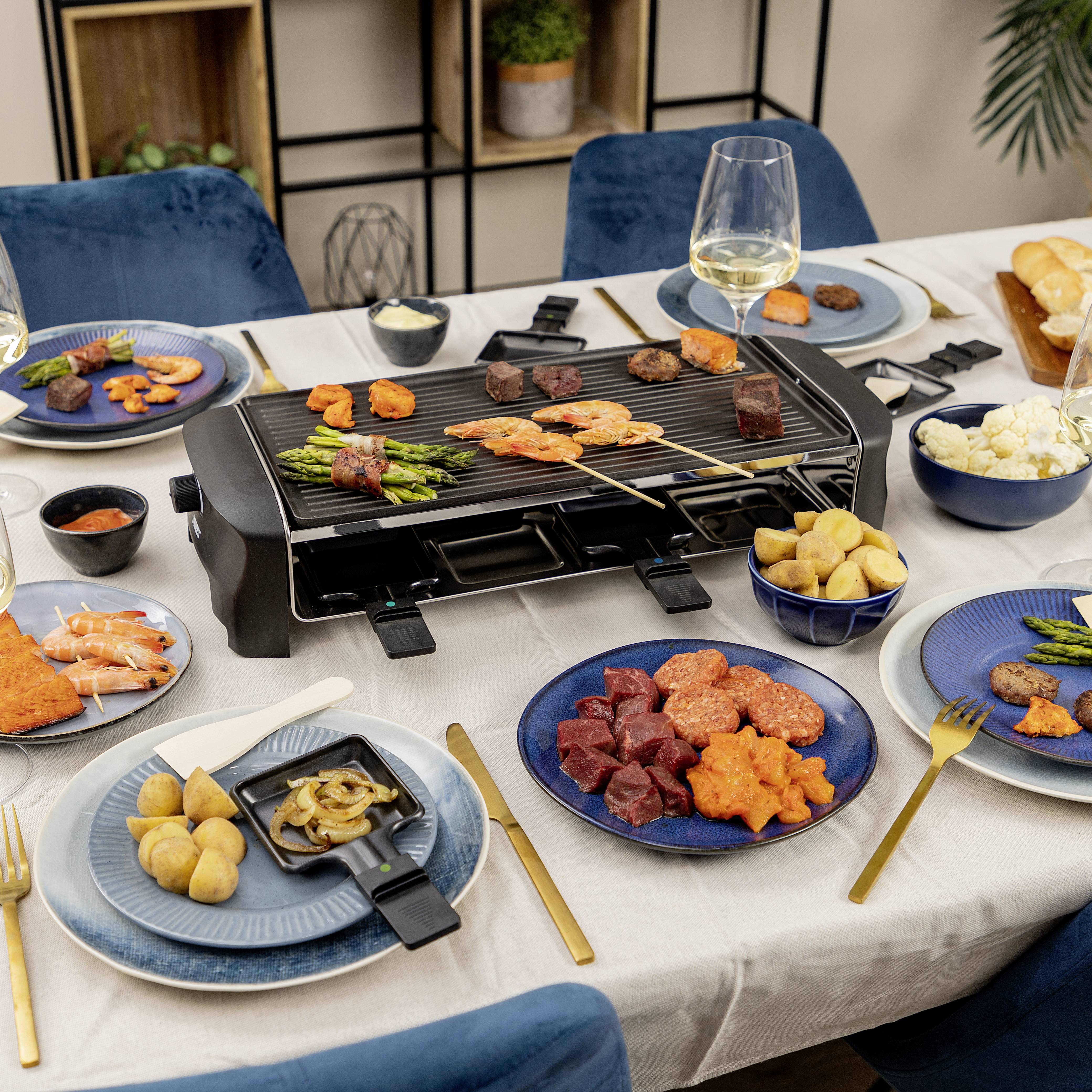 Ein gedeckter Tisch mit einem Tischgrill in der Mitte, umgeben von Tellern mit Fleisch, Gemüse, Soßen und gefüllten Gläsern.
