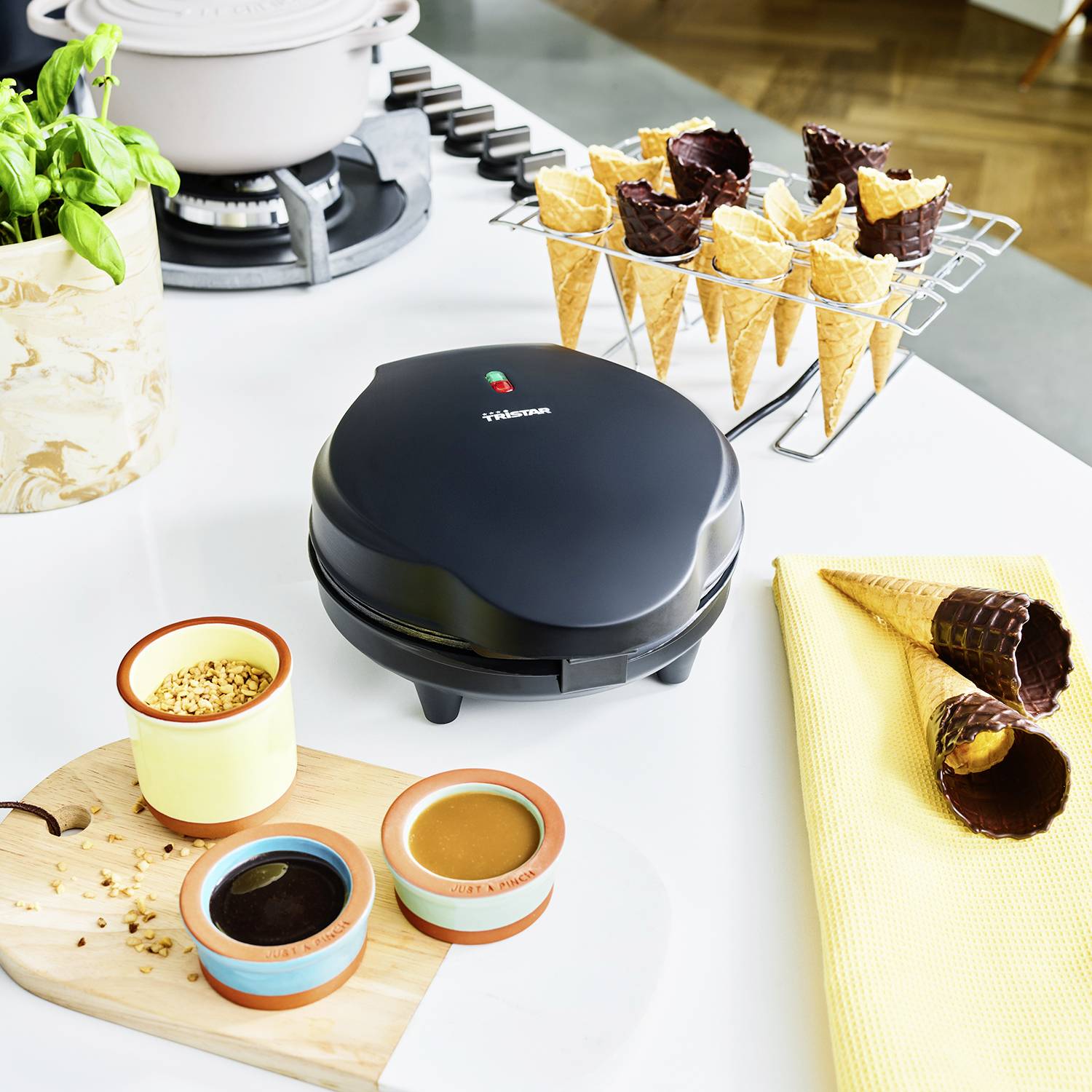 Waffel-Maker auf Küchenarbeitsplatte, umgeben von Waffelhörnchen und Soßen. Basilikum in Vase, Kochtopf auf Herd im Hintergrund.