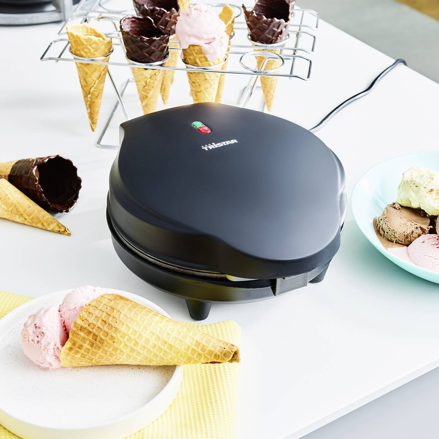 Ein Waffeleisen mit mehreren Eiswaffeln in Halterung auf Tisch. Daneben eine Schale mit drei Kugeln Eis und ein Teller mit Eistüte.