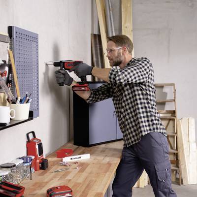 Ein Mann benutzt eine Bohrmaschine, um ein Loch in eine Wand zu bohren. Neben ihm ist ein Tisch mit Werkzeugen und eine Leiter zu sehen.