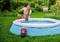 Eine Frau pumpt mit einem elektrischen Pumpgerät Wasser in einen aufblasbaren Pool im Garten. Im Hintergrund ist viel Grün zu sehen.