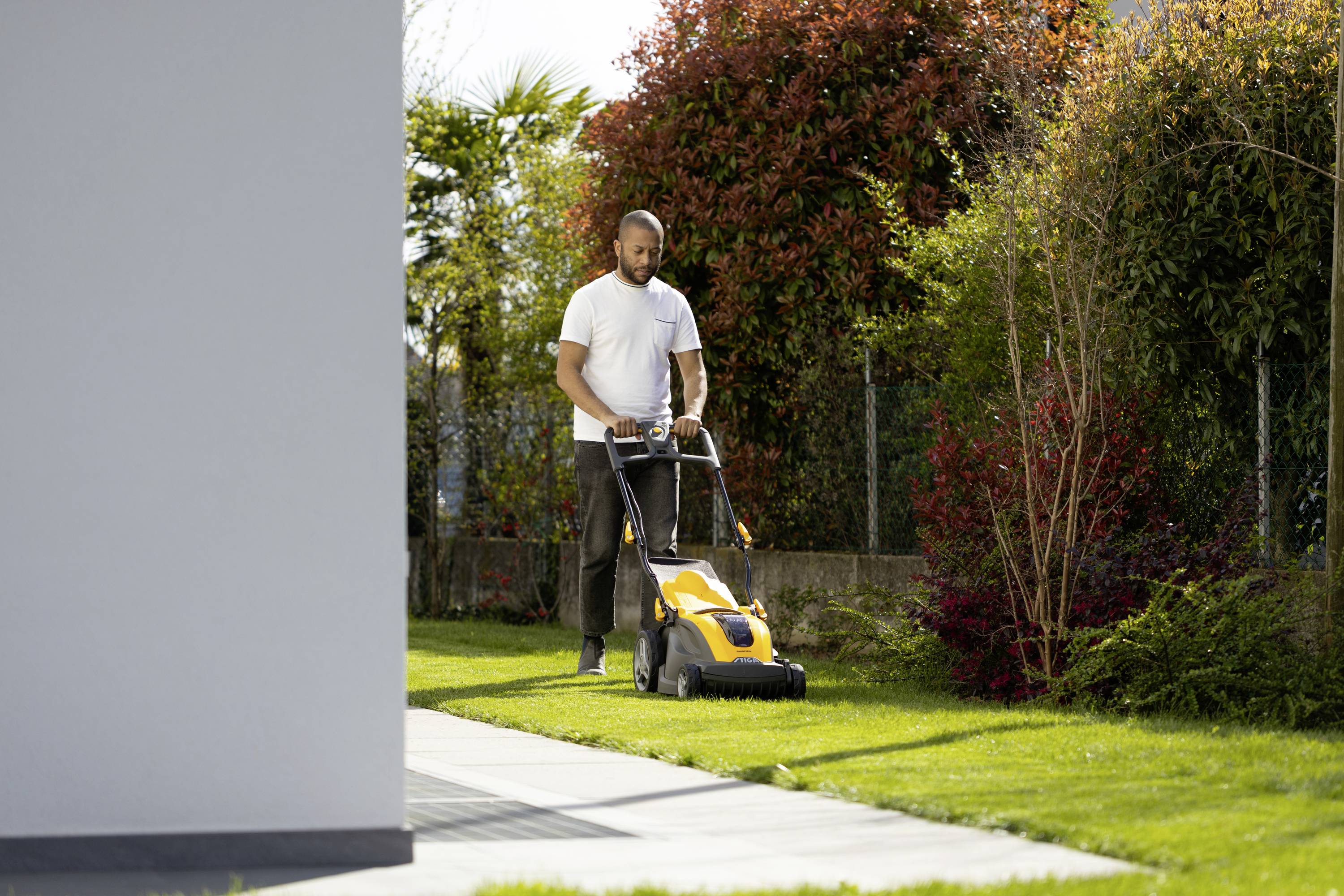 Ein Mann mäht den Rasen mit einem elektrischen Rasenmäher in einem Garten. Im Hintergrund sind Sträucher und Bäume zu sehen.