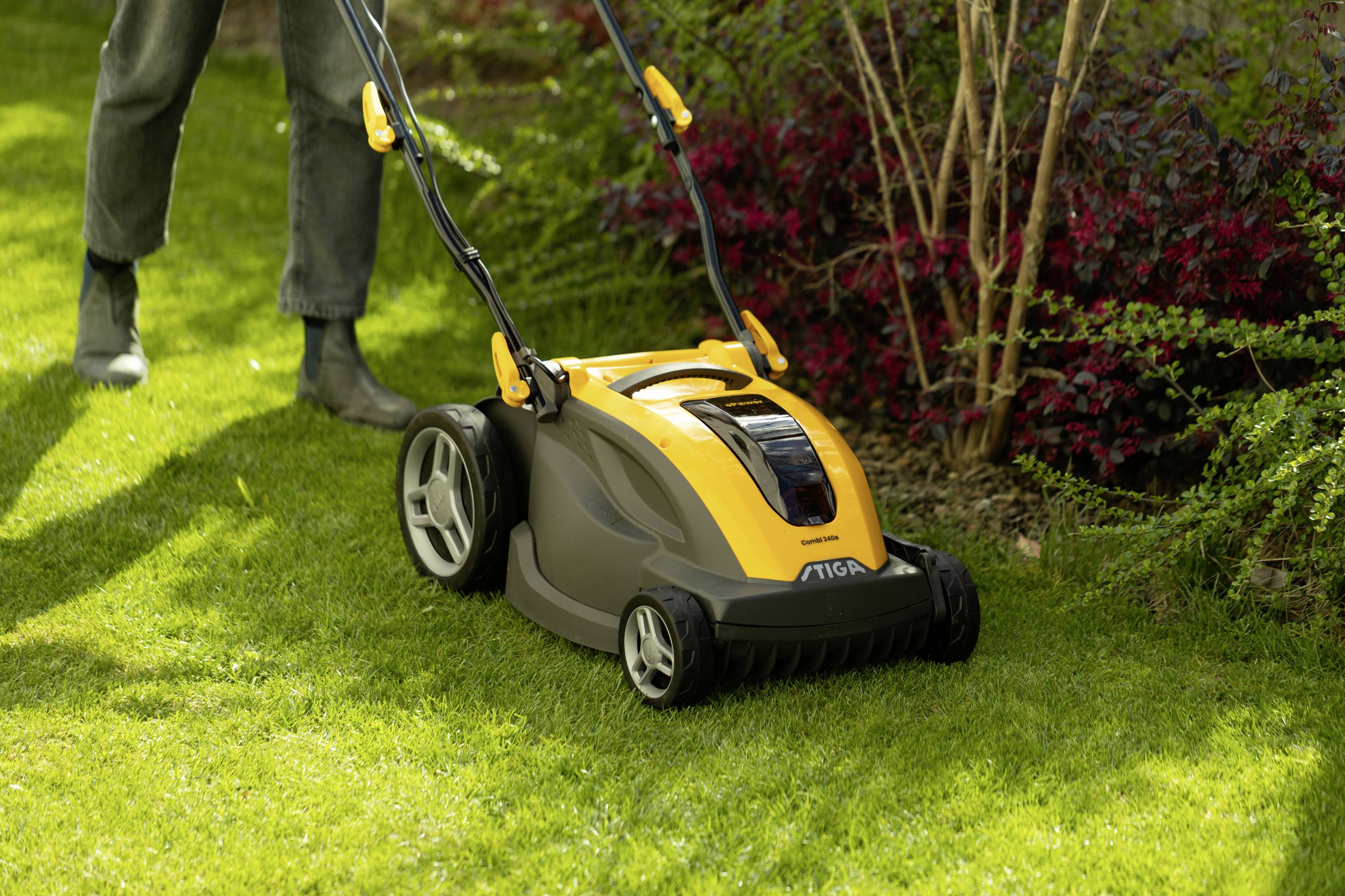 Eine Person mäht mit einem gelben Rasenmäher das Gras im Garten. Rechts sind Büsche zu sehen, die den Rasenbereich säumen.