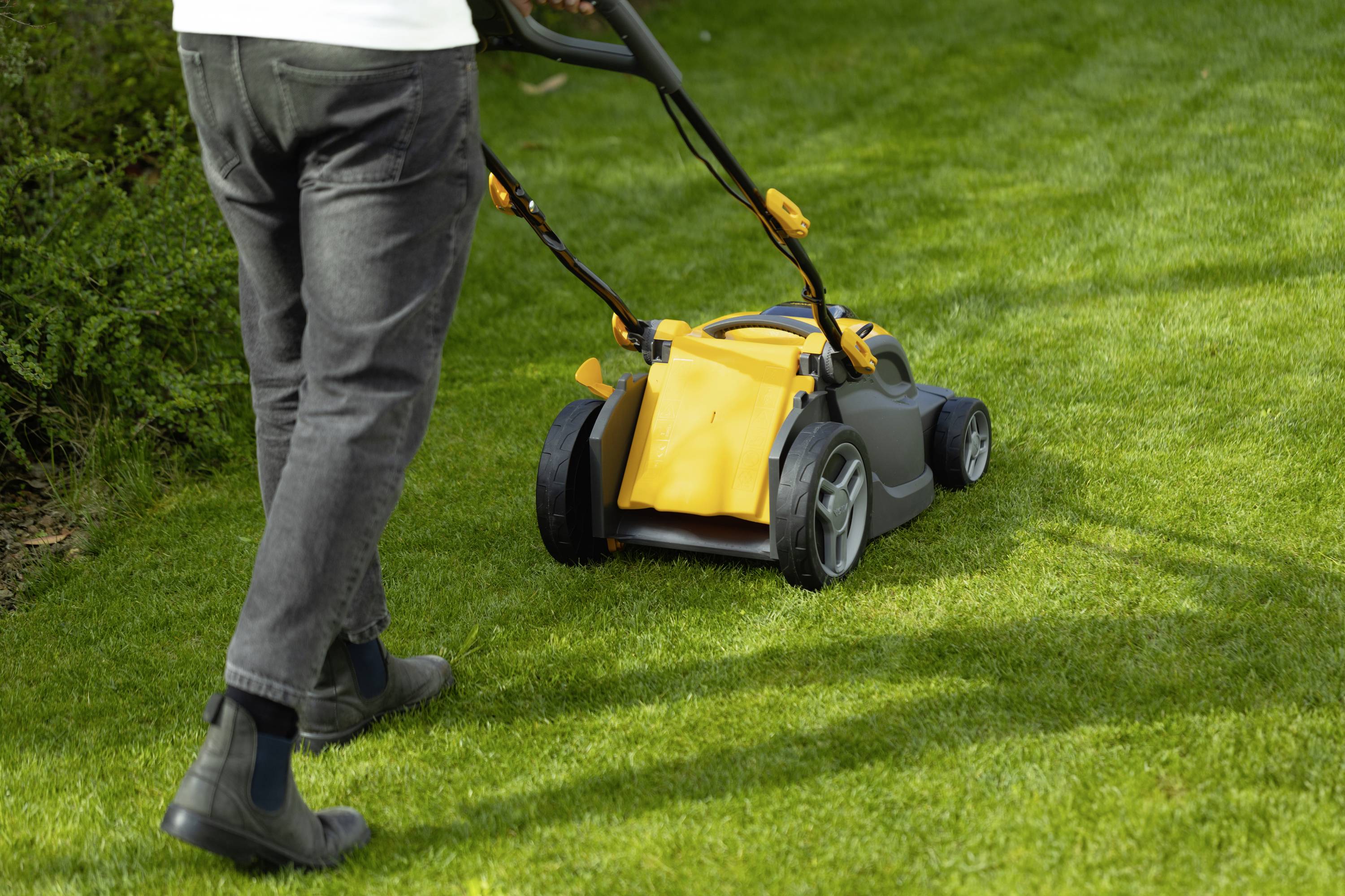 Person mäht Rasen mit einem gelben Rasenmäher. Sicht von hinten, die Person ist in schwarze Kleidung gekleidet. Gras ist grün und gepflegt.