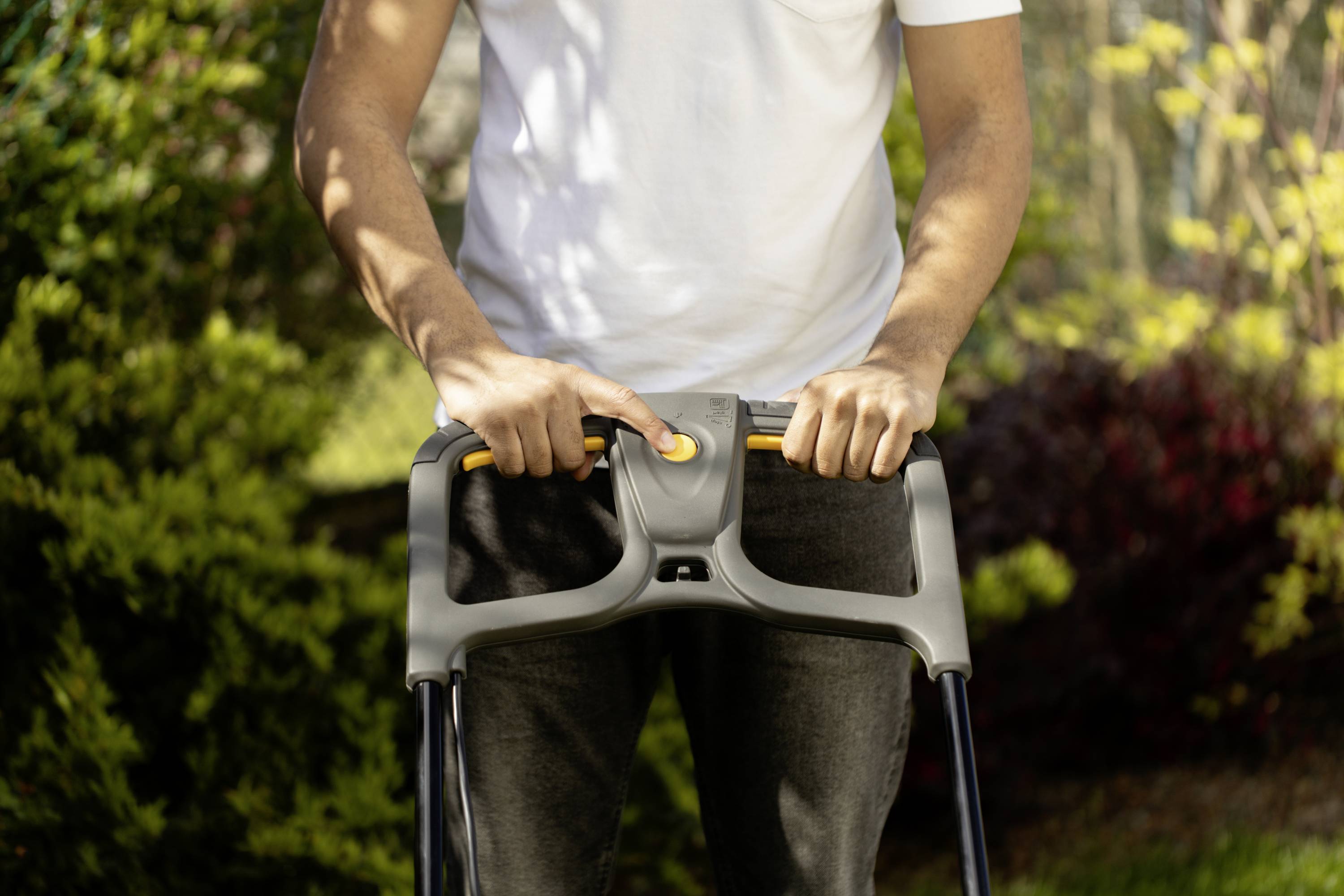 Person schiebt Rollator in einem Garten bei Tageslicht, umgeben von grünen Pflanzen und Büschen. Der Fokus liegt auf den Händen.