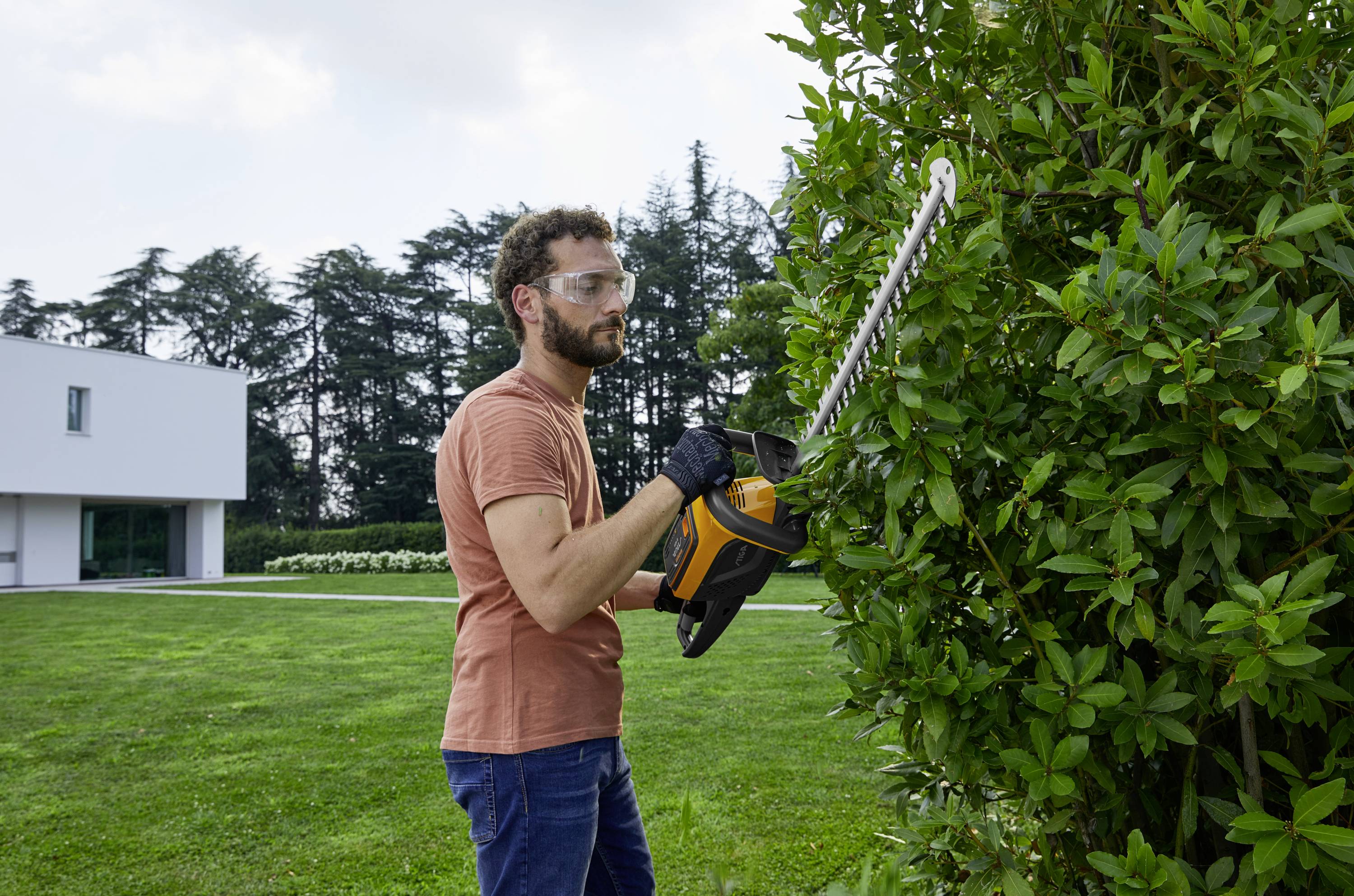 Ein Mann schneidet mit einer elektrischen Heckenschere eine Hecke in einem Garten. Im Hintergrund ist ein modernes Gebäude zu sehen.