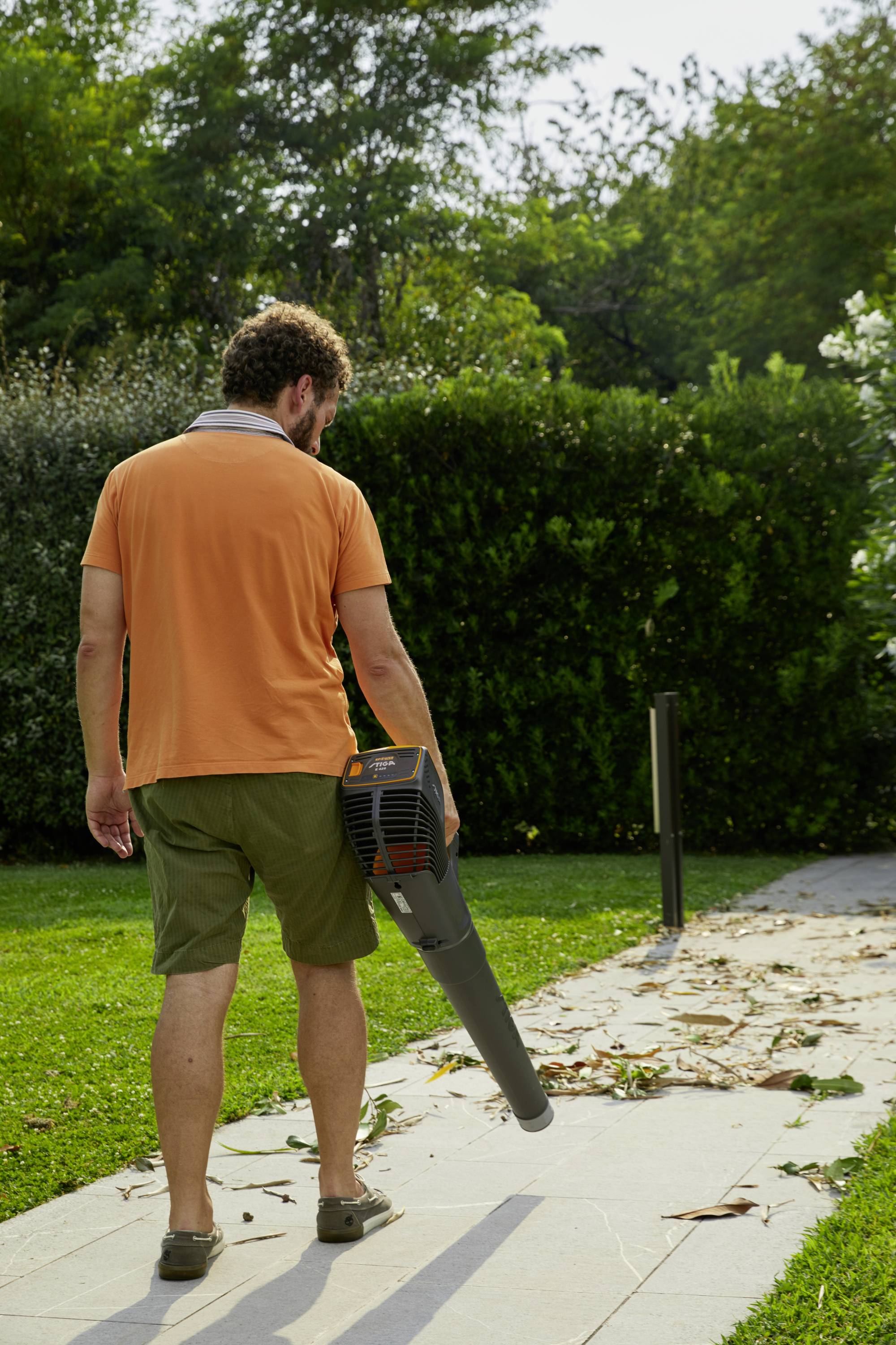 Ein Mann benutzt einen Laubbläser, um Laub auf einem Gehweg in einem Garten zu entfernen. Im Hintergrund sind Sträucher zu sehen.