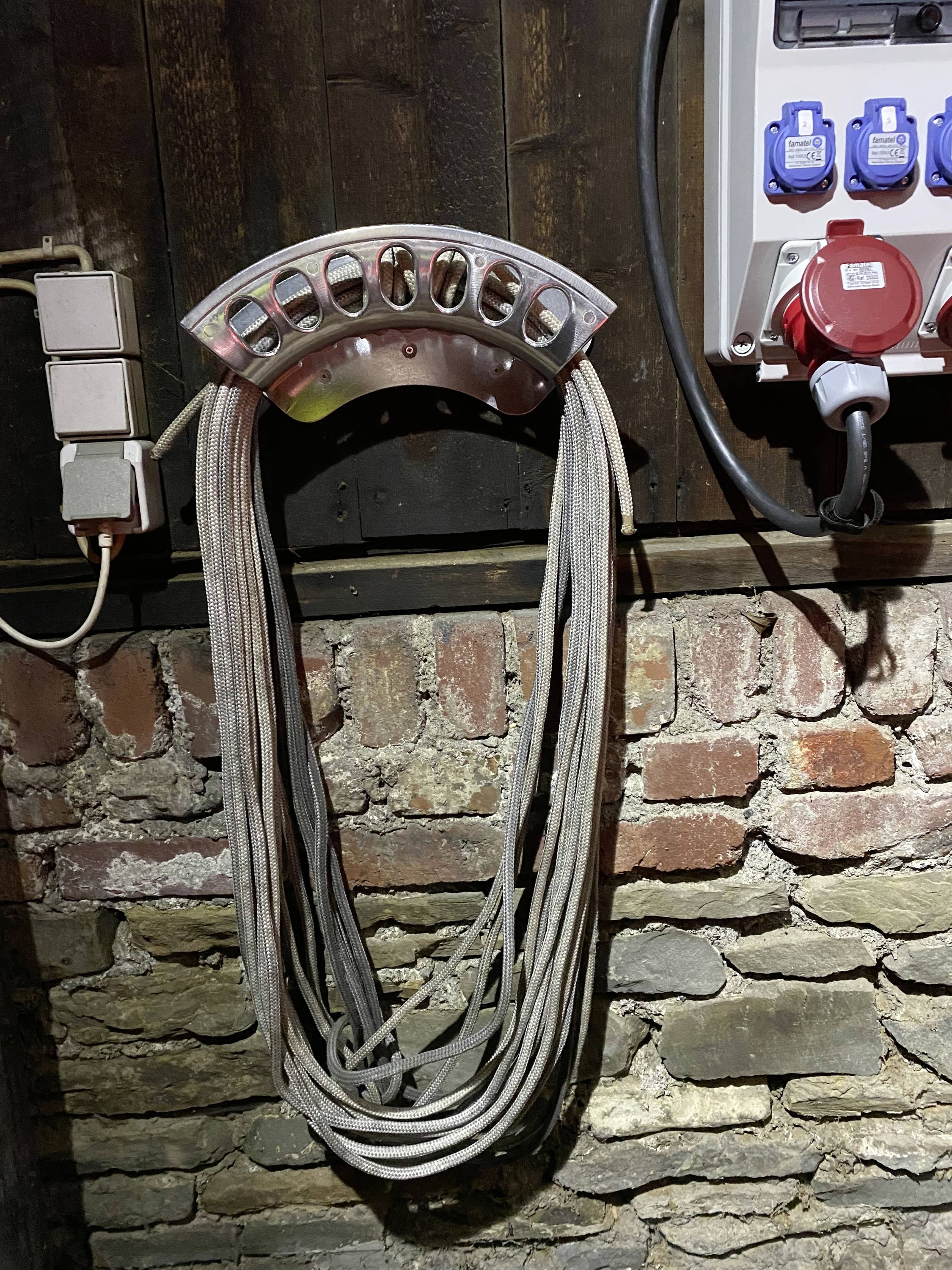 Ein Bündel Kabel hängt an einer Halterung an einer Ziegelwand. Daneben sind Steckdosen und ein elektrischer Anschluss sichtbar.