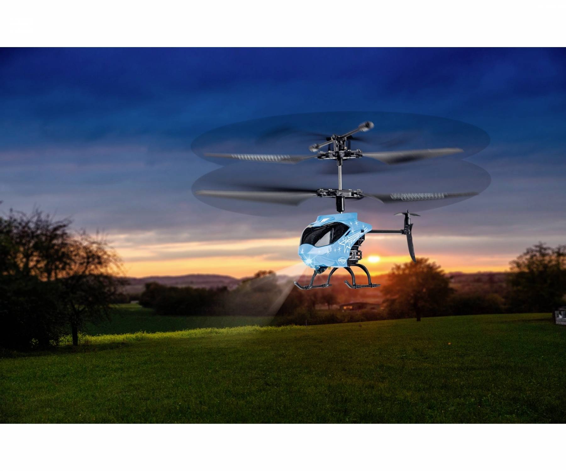 Ein blaues Spielzeug-Helikopter-Modell schwebt während eines Sonnenuntergangs über einer grünen Landschaft mit Bäumen im Hintergrund.