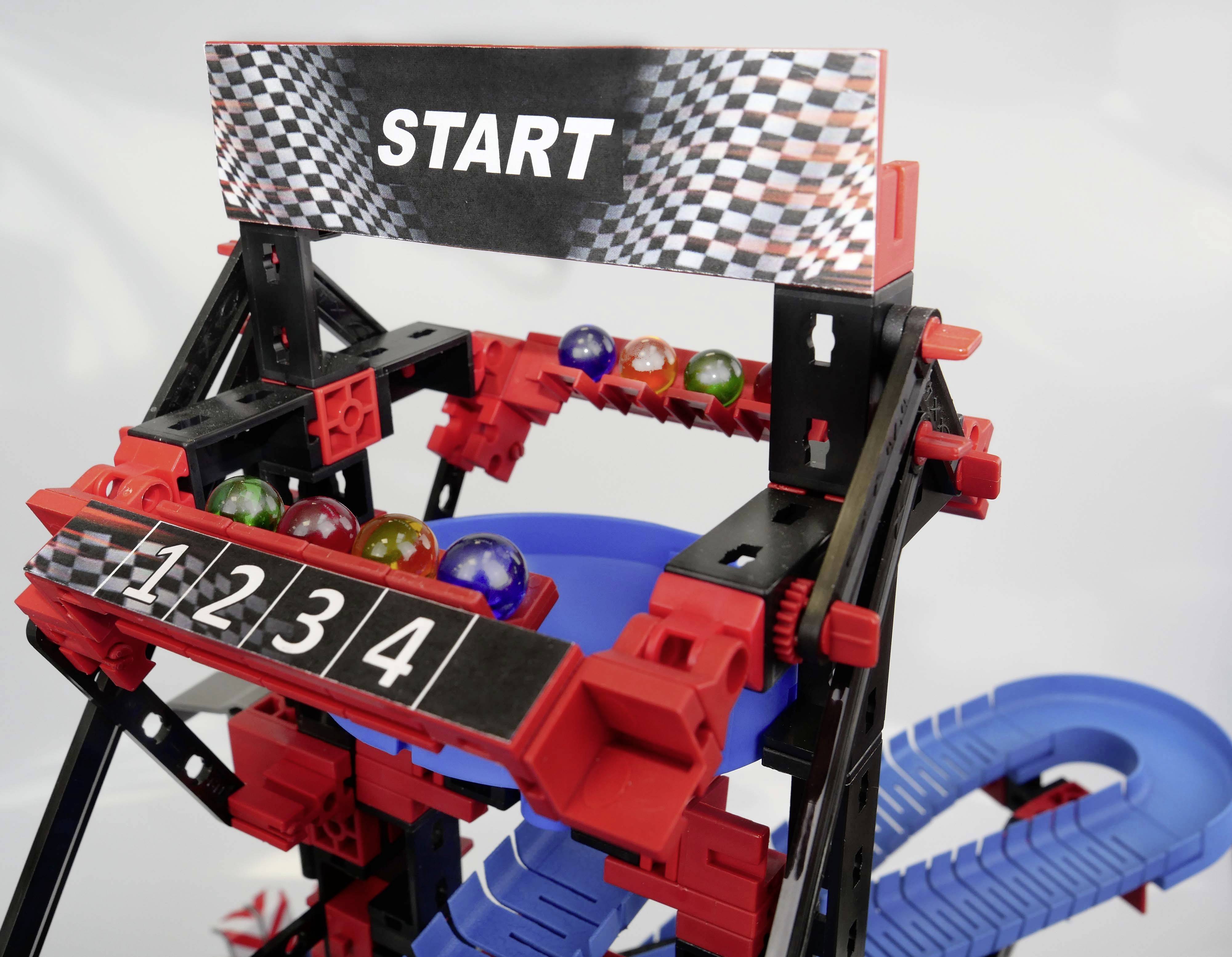 Startlinie eines Murmelrennens mit vier nummerierten Bahnen und bunten Murmeln. Ein schwarzes Schild mit 'START'.