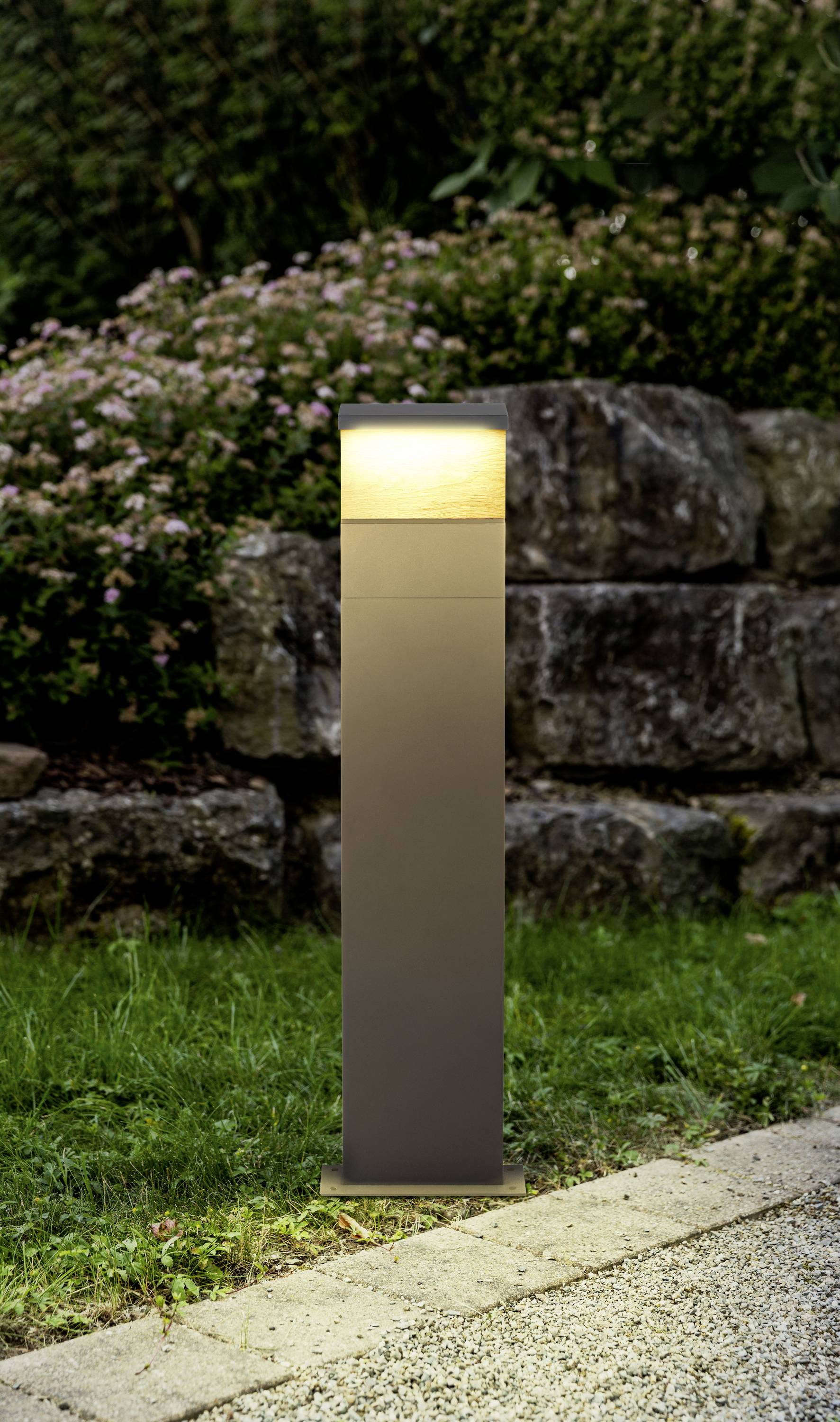 Eine moderne Gartenlaterne leuchtet auf einem Rasen neben einem gepflasterten Weg, umgeben von Grünpflanzen und einer Steinmauer.