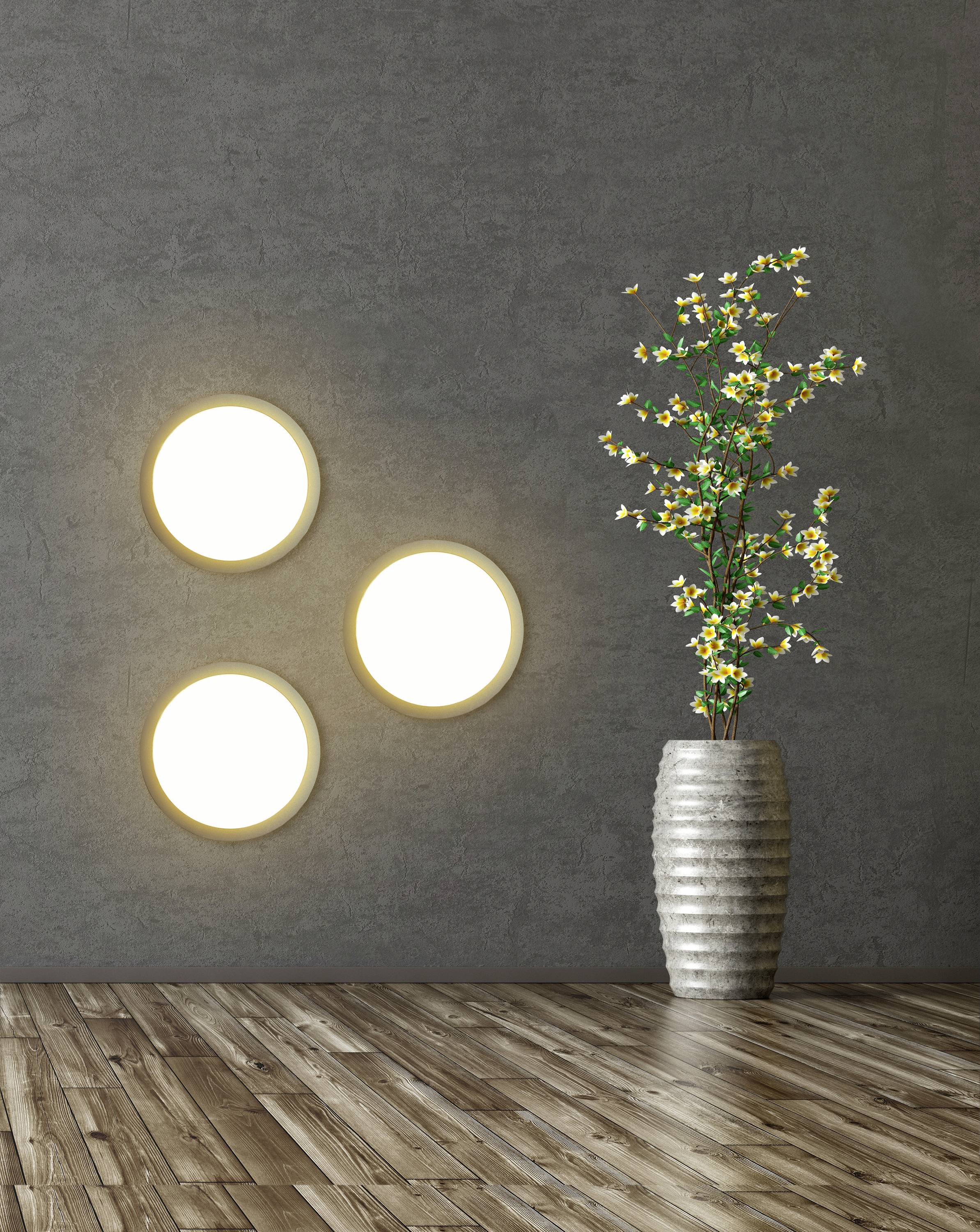 Moderner Innenraum mit grauer Wand, drei runden leuchtenden Wandlampen und einer hohen Vase mit gelb blühenden Zweigen auf Holzboden.