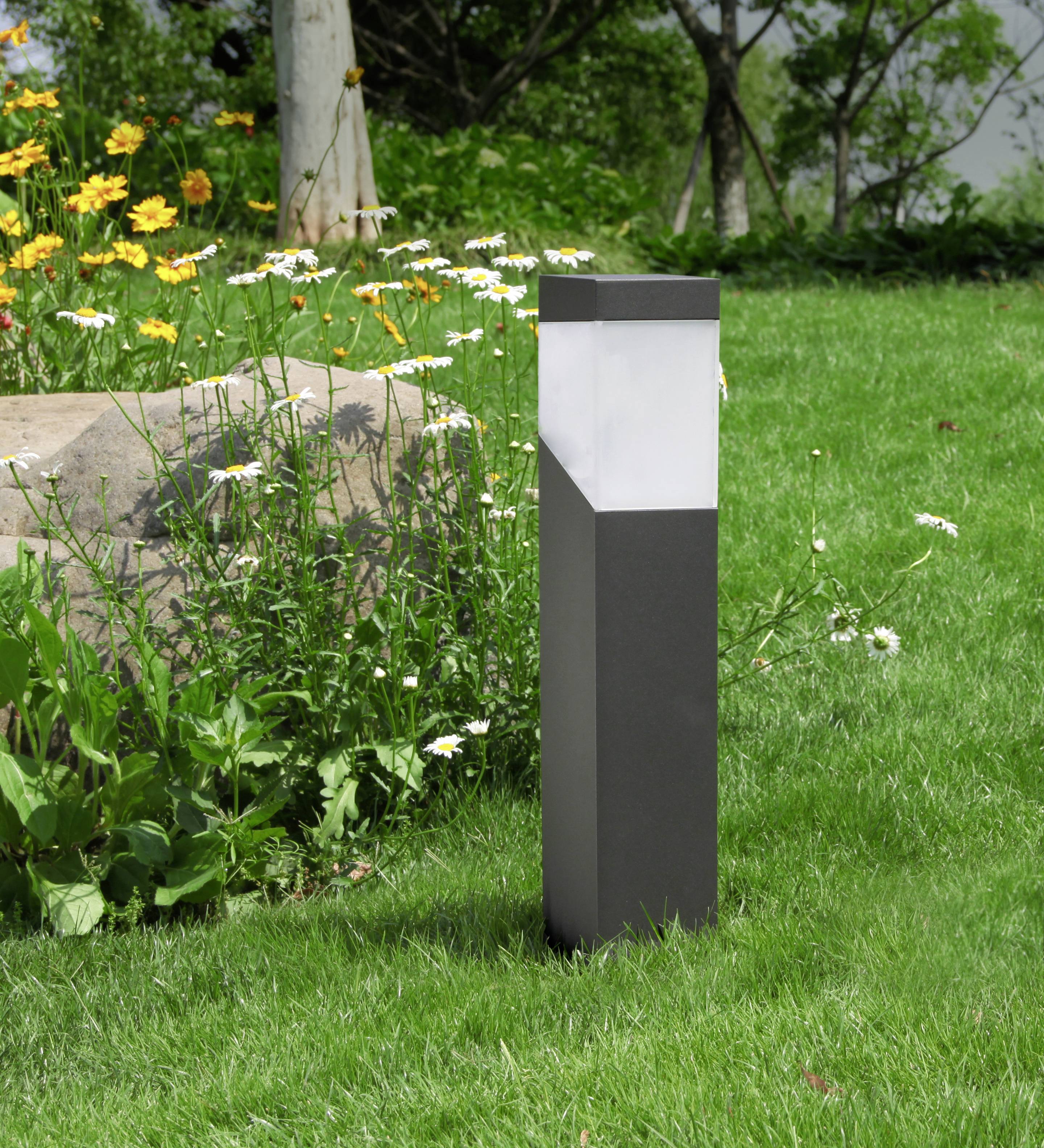 Eine moderne Gartenlampe aus Metall steht auf einem grünen Rasen, umgeben von gelben und weißen Blumen sowie einem großen Stein.
