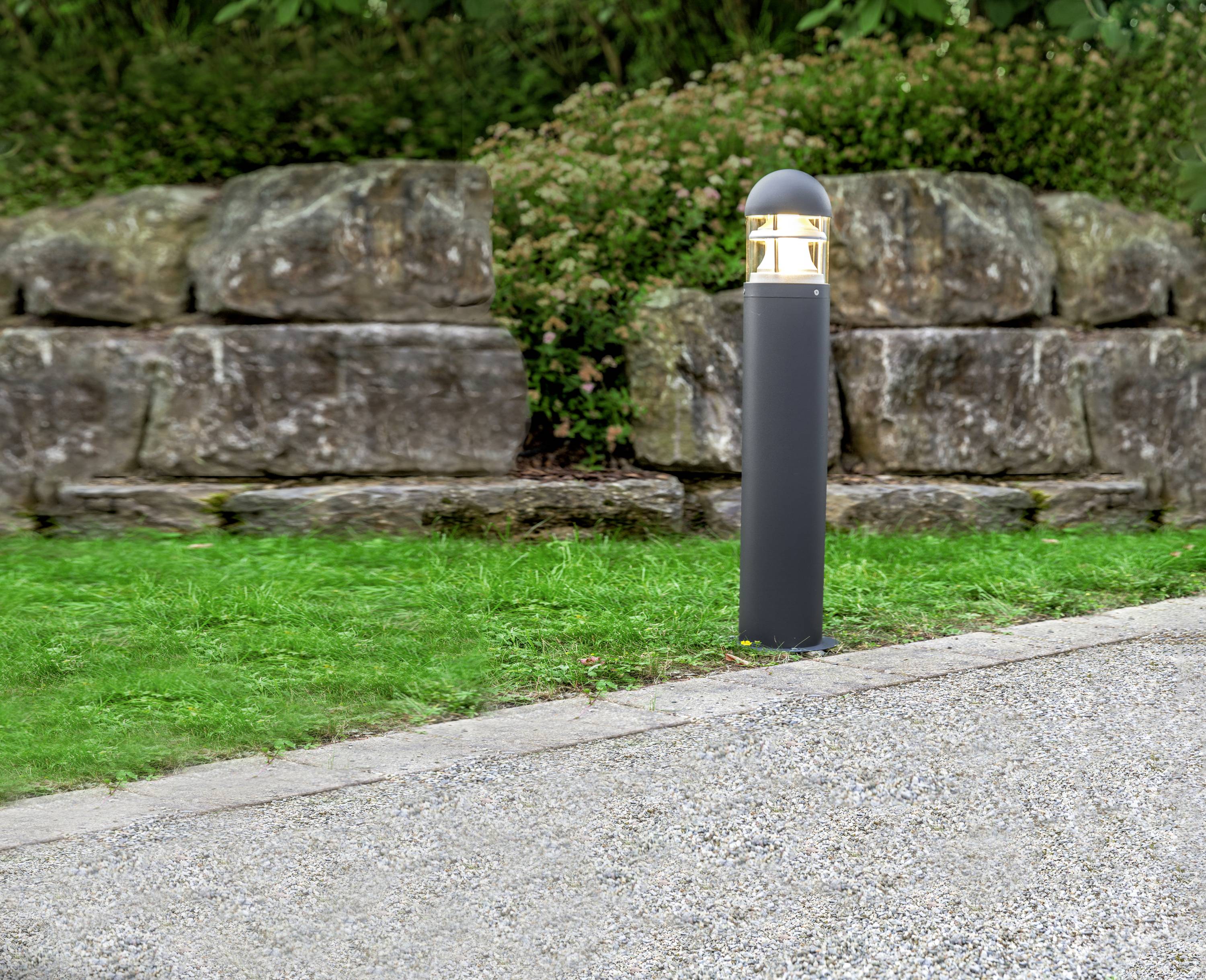 Ein beleuchteter Wegweiser steht neben einem gepflasterten Weg, umgeben von Gras und einer niedrigen Steinmauer im Hintergrund.