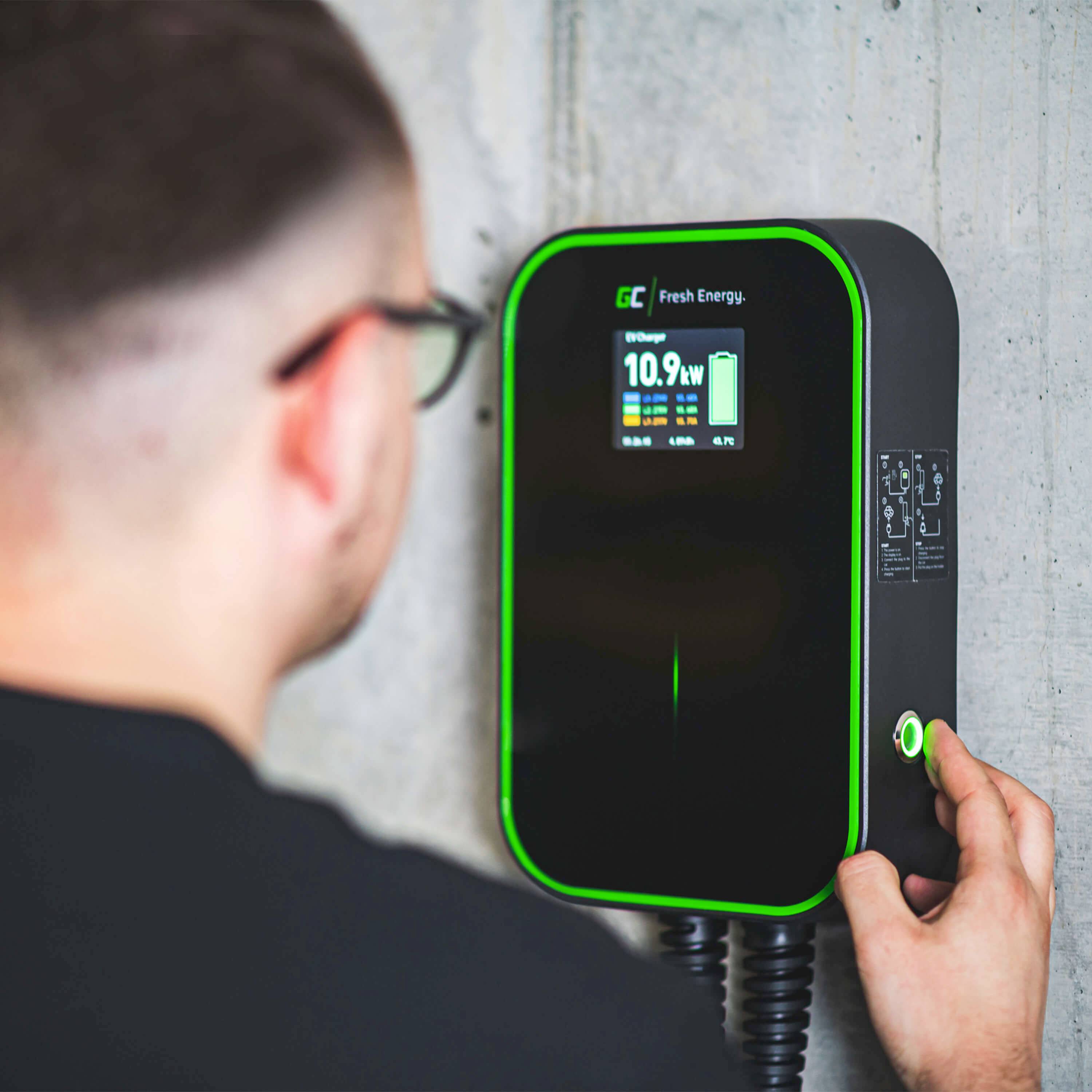 Ein Mann mit Brille benutzt eine Wandladestation für Elektrofahrzeuge. Das Display zeigt 'Fresh Energy' und einen Stromverbrauch von 10,9 kW an.