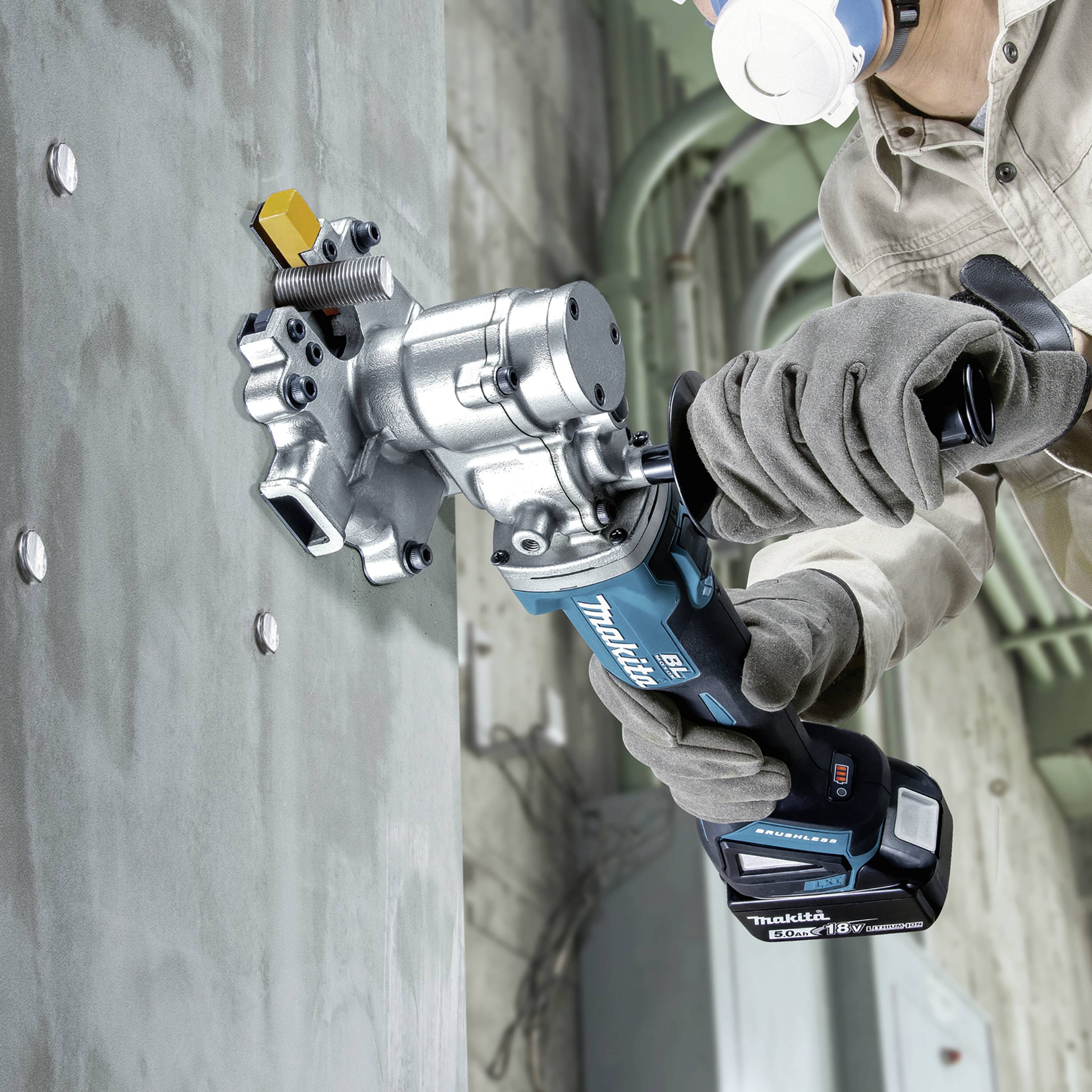 Ein Arbeiter trägt eine Schutzmaske und Handschuhe und nutzt eine blaue elektrische Bohrmaschine, um ein Metallstück an einer Betonwand zu befestigen.