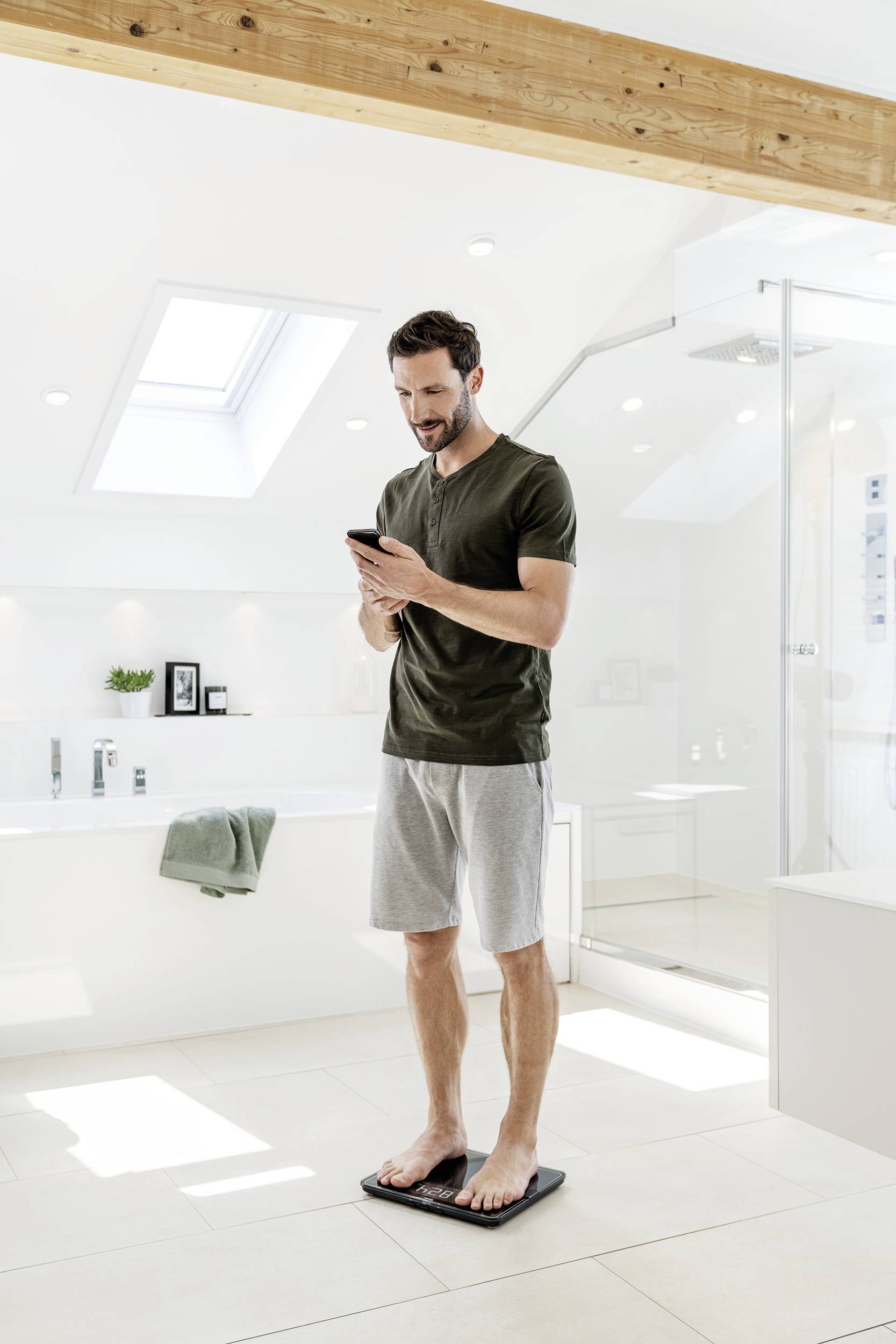 Ein Mann steht in einem Badezimmer auf einer Waage und schaut auf sein Smartphone.