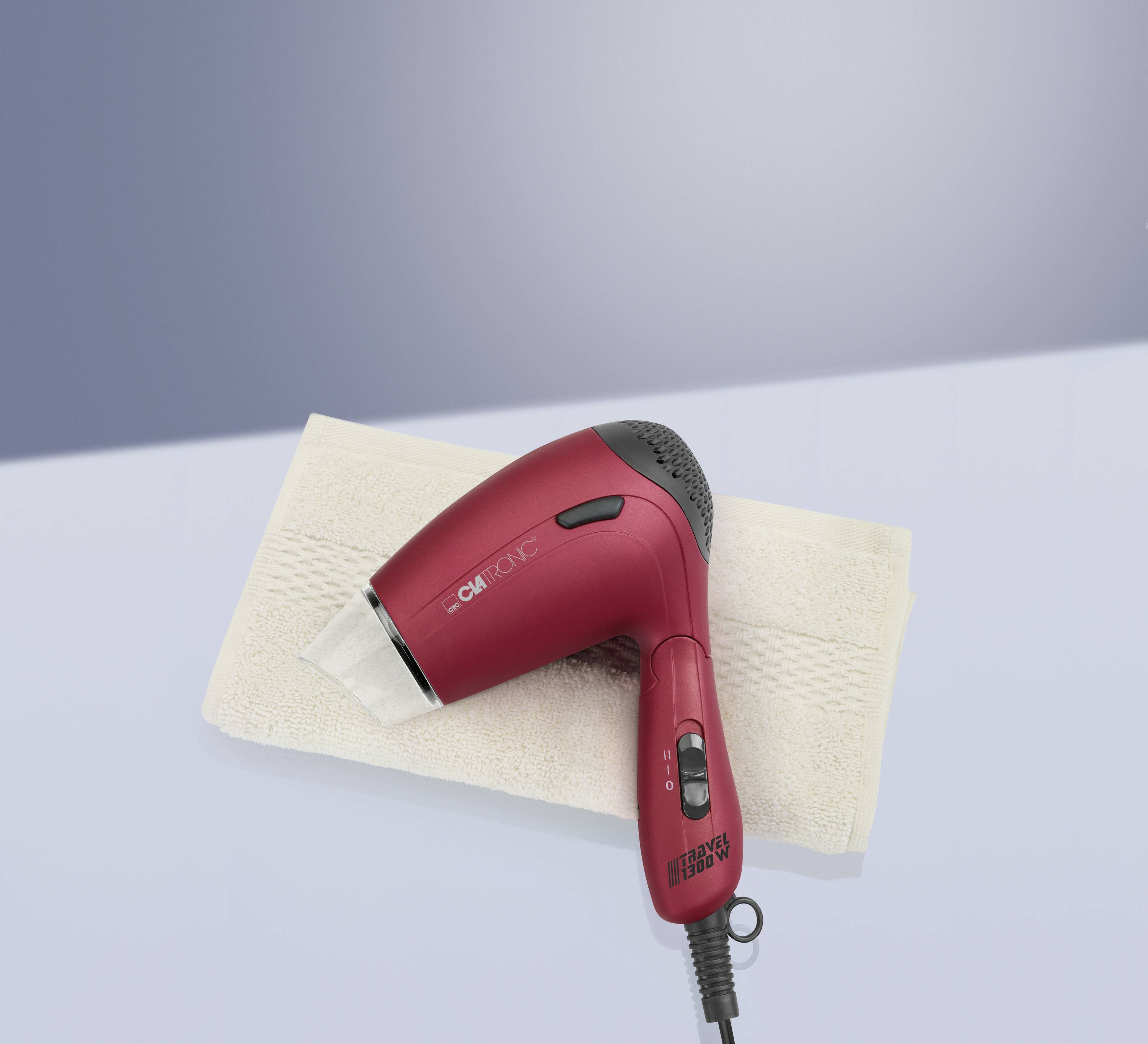 Ein roter Haartrockner liegt auf einem weißen Handtuch. Der Haartrockner ist eingeschaltet und liegt auf einer glatten Oberfläche.