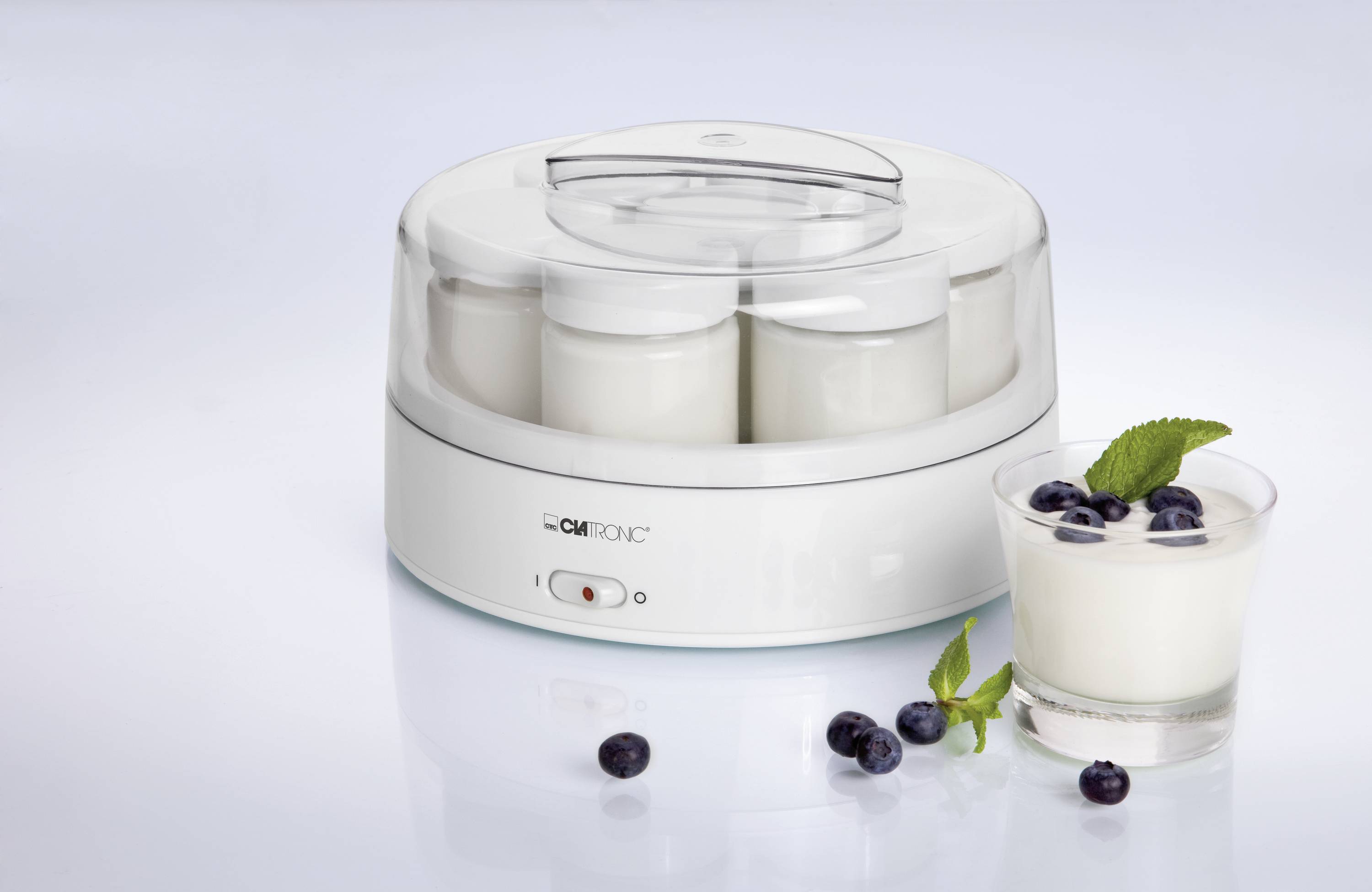Ein elektrischer Joghurtbereiter mit mehreren Gläsern gefüllt mit Joghurt. Daneben ein Glas Joghurt mit Blaubeeren und Minze.