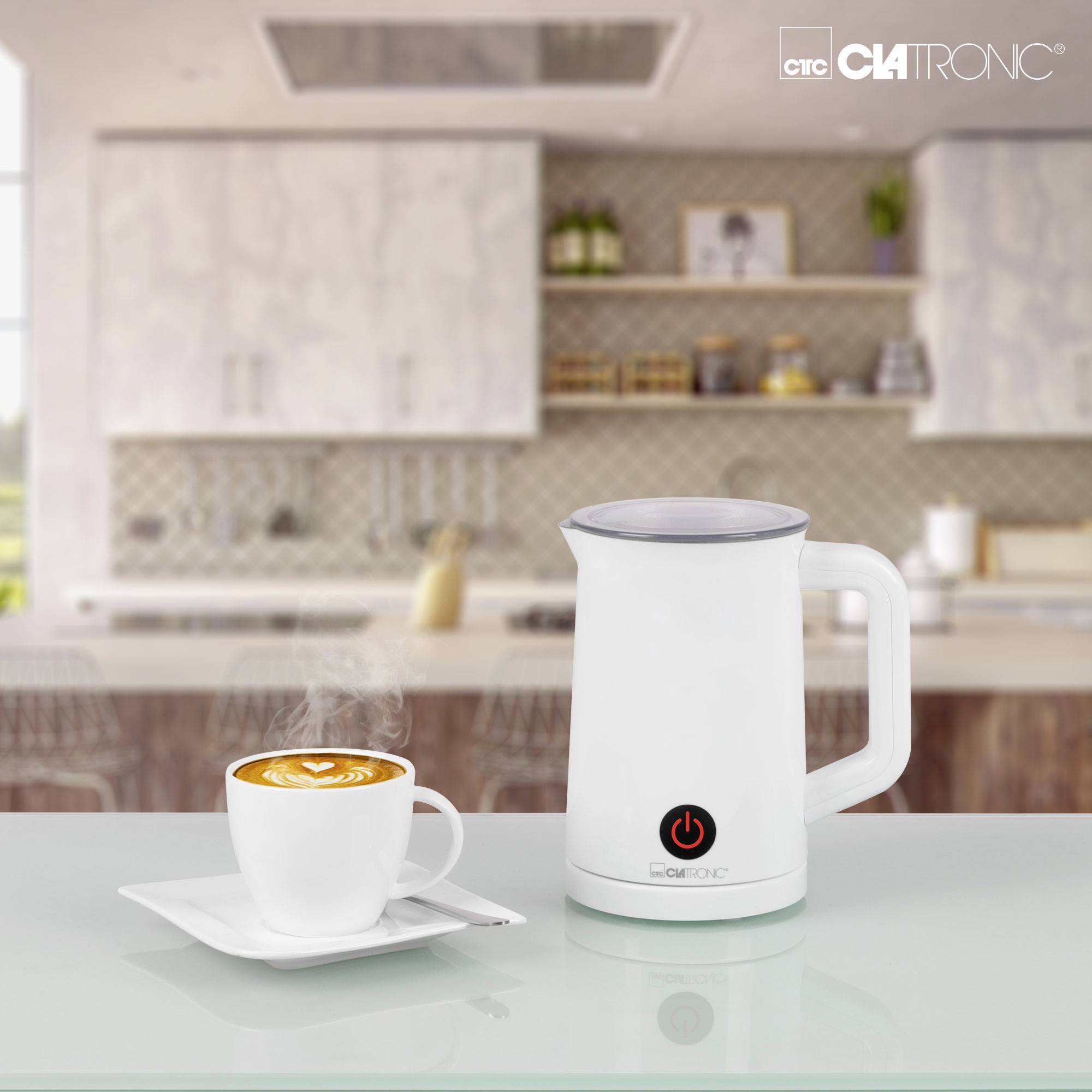 Ein weißer Milchaufschäumer und eine Tasse Kaffee mit Latte Art stehen auf einem Tisch in einer modernen Küche im Hintergrund.