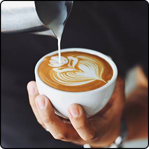 Eine Person gießt kunstvoll geschäumte Milch in eine Tasse Kaffee, um ein Herzmuster zu kreieren.